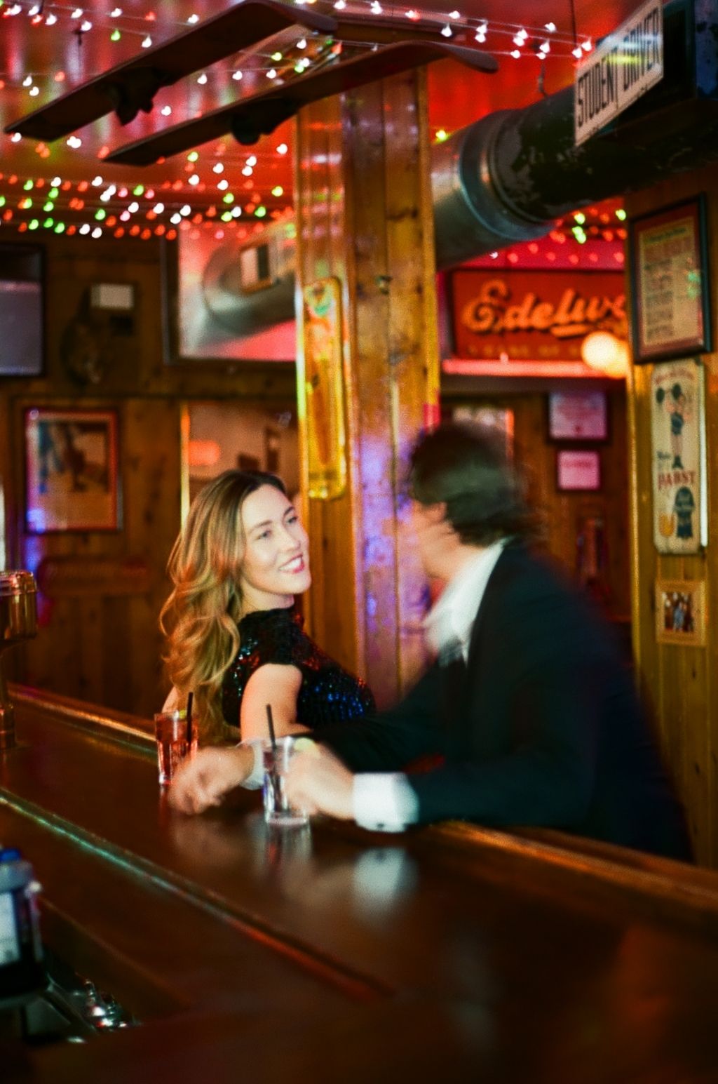 A smiling woman with long hair sits at a warmly lit wooden bar adorned with colorful string lights.