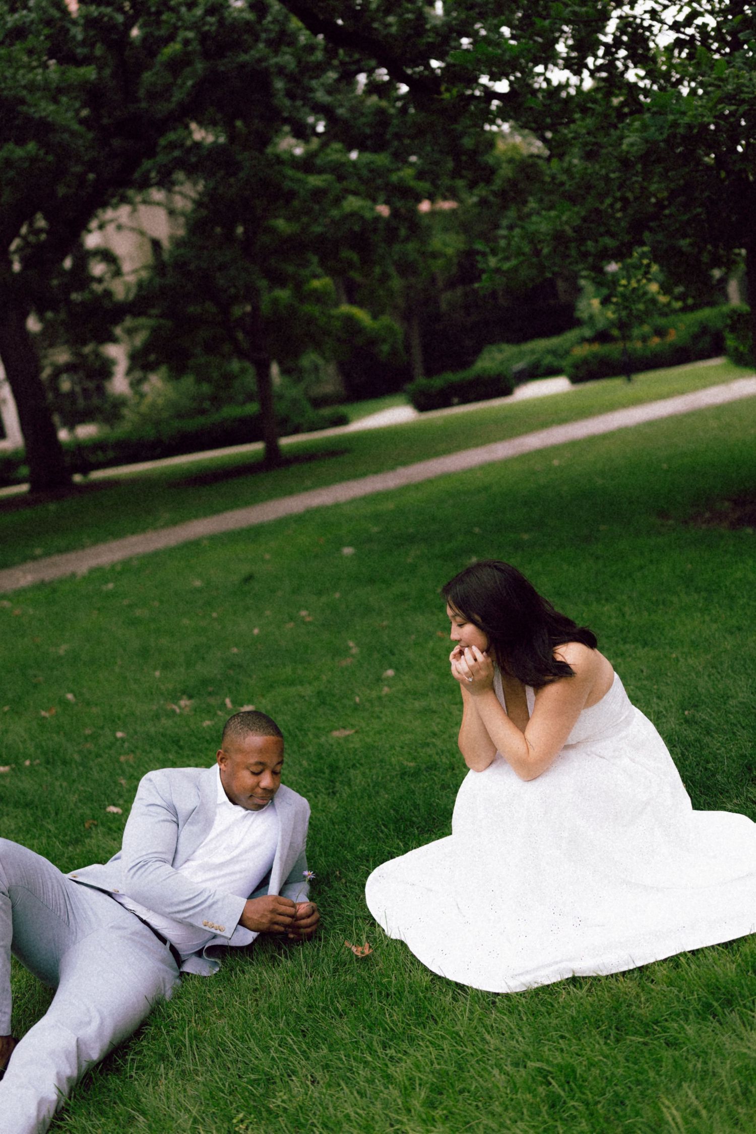 Interracial couple in wedding attire sitting on green grass in a park, bride in white dress and groom in gray suit.