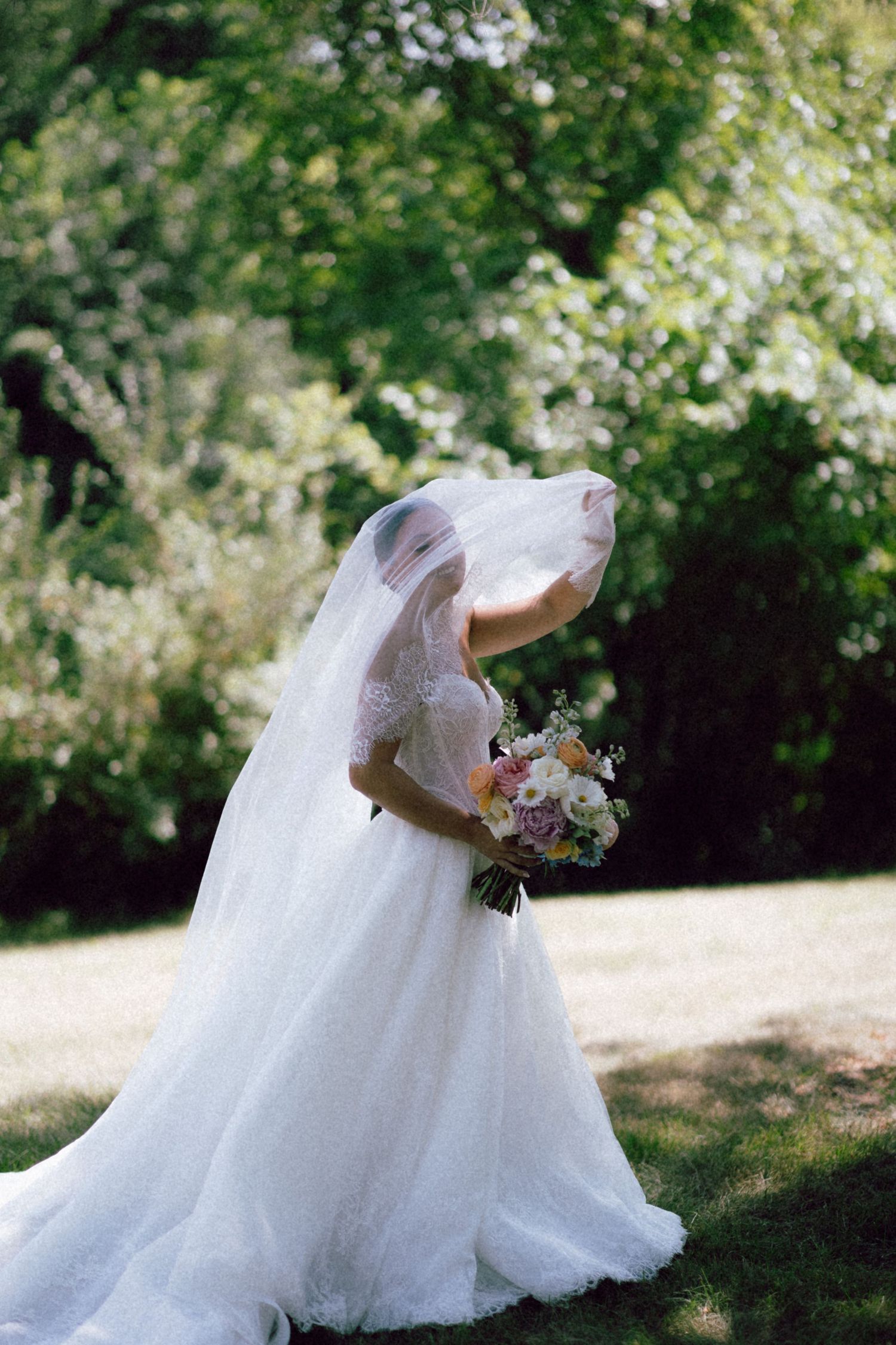 Bride in white ball gown with veil blowing over face holds colorful bouquet outdoors in lush green garden.