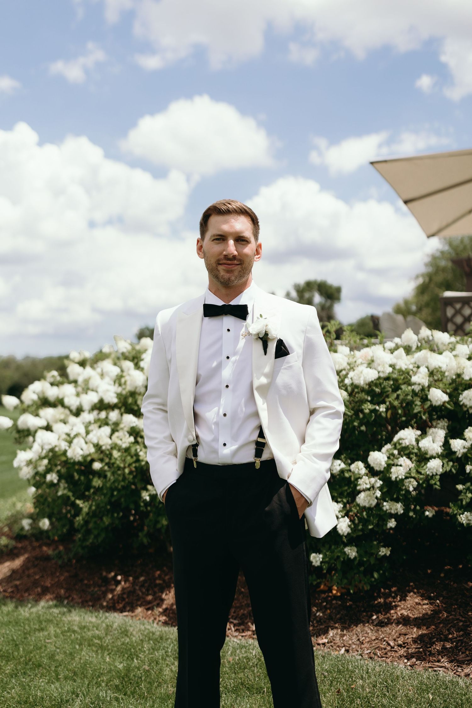 A dapper groom in a white dinner jacket poses in front of blooming white flowers on a sunny day.