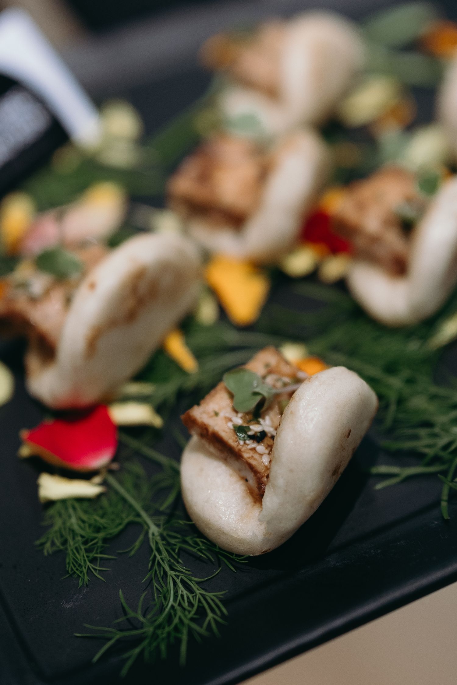 Gourmet bao buns garnished with herbs and flower petals on a black plate.
