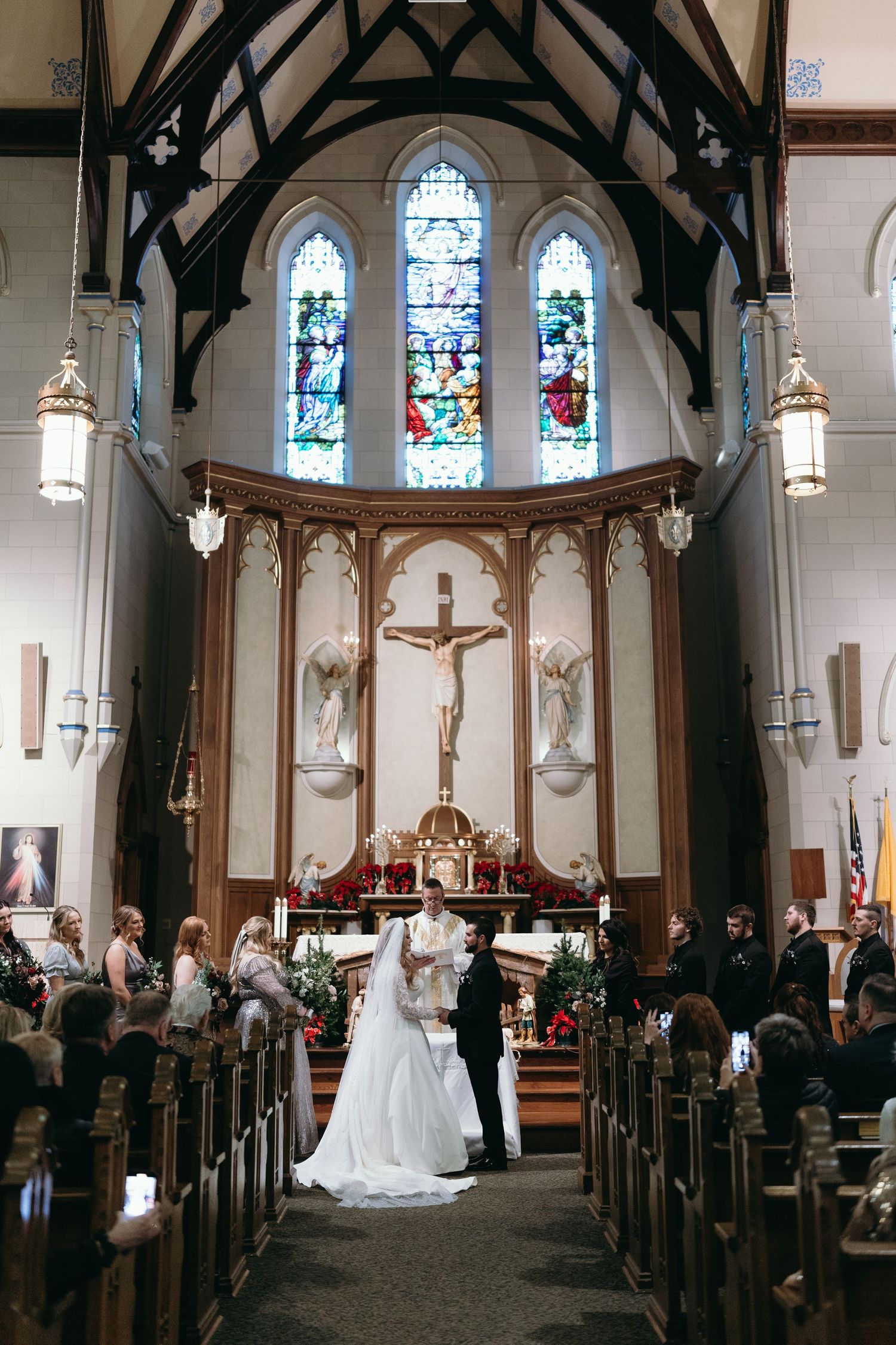 Wedding ceremony taking place beneath stained glass windows and crucifix in ornate church sanctuary.