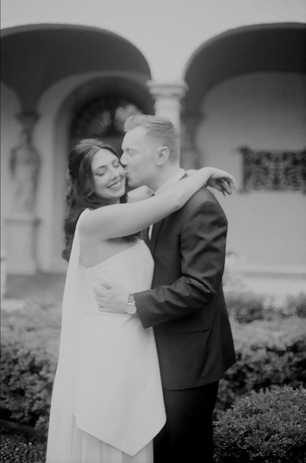 A tender black and white moment between a couple embracing during a wedding photoshoot in front of arched architecture.