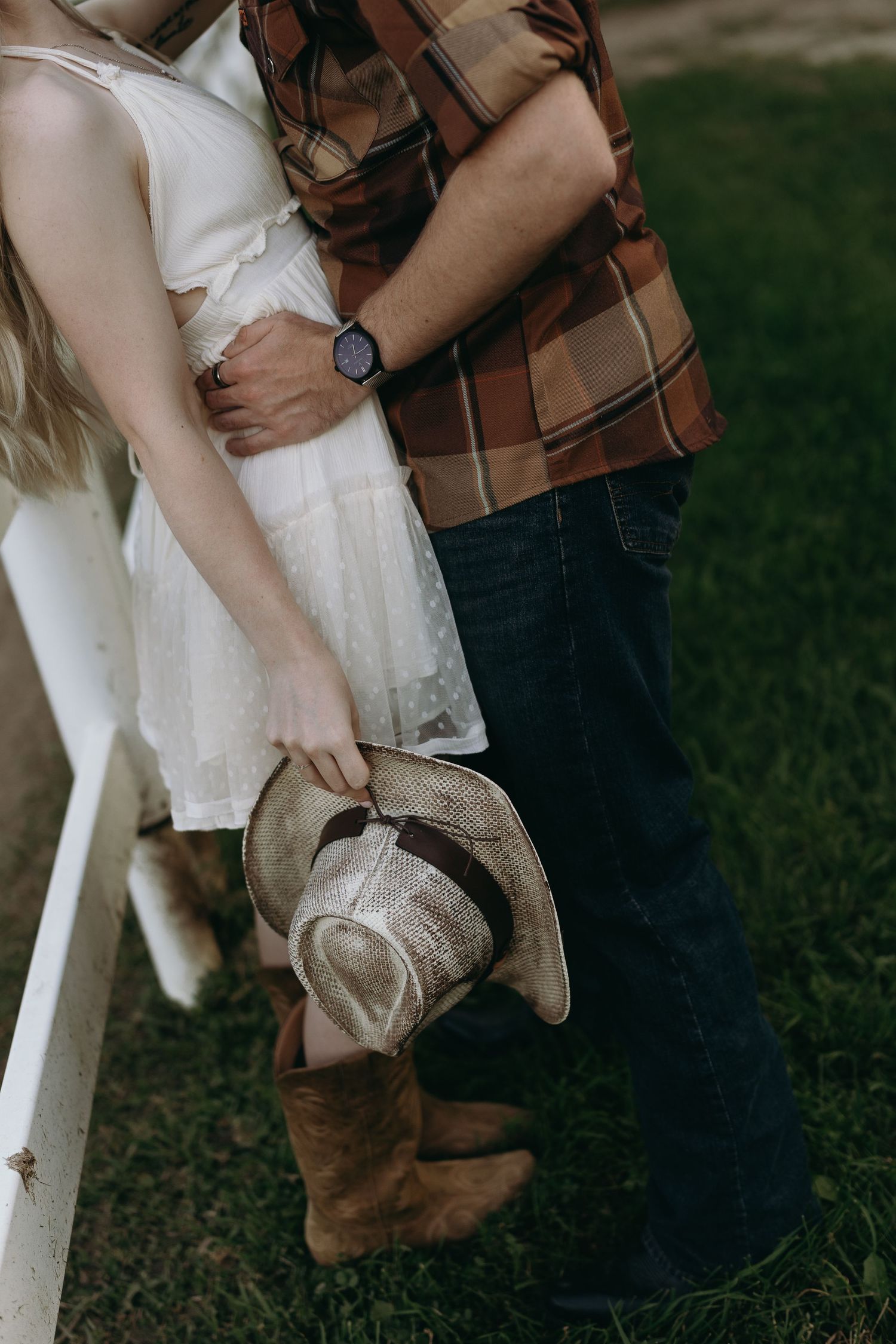 A couple holds hands while wearing a brown jacket and white dress in an outdoor setting near grass.