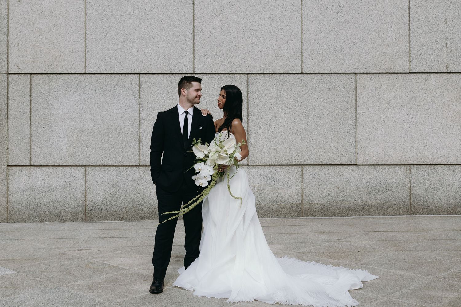 A couple in wedding attire embraces in front of a modern concrete wall with a white floral bouquet.