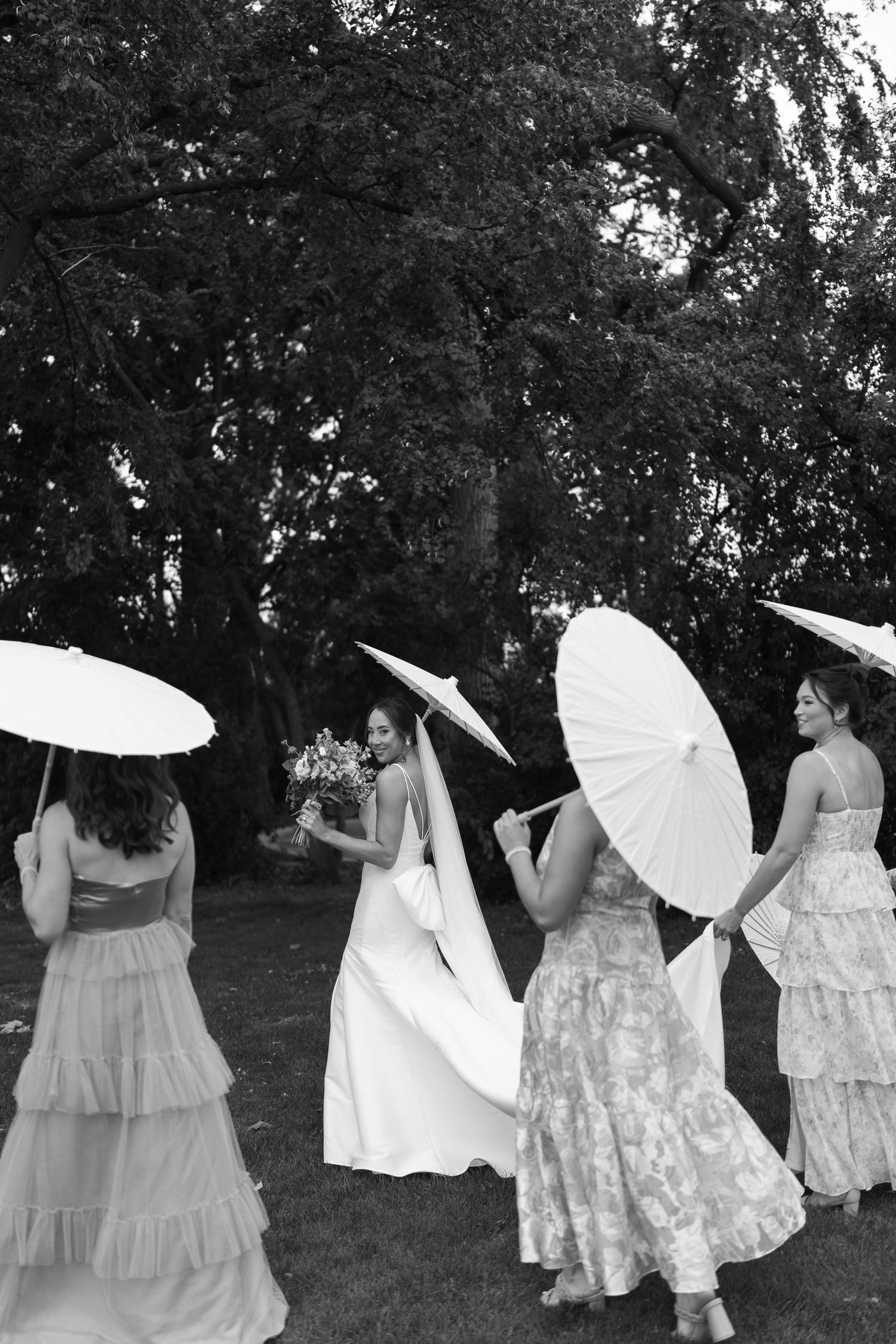 Black and white photo of bridal party with parasols under trees.