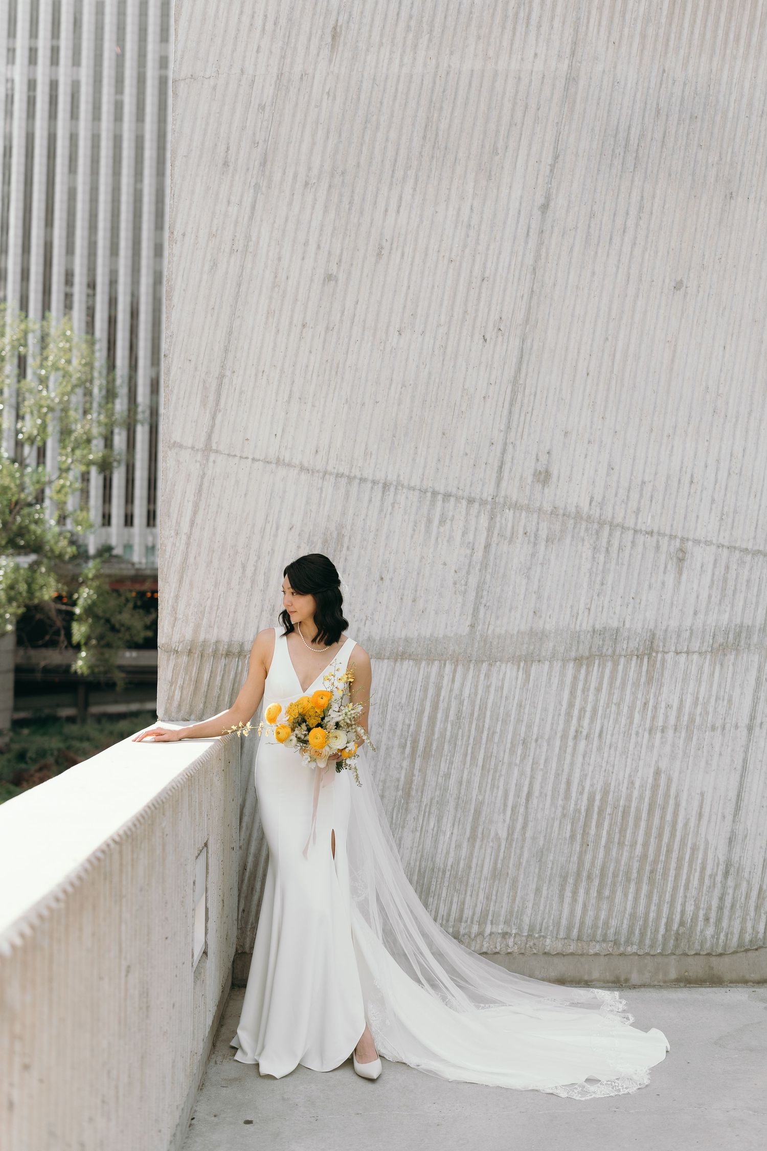 Minimalist bridal portrait against textured white wall with flowing dress and yellow bouquet.