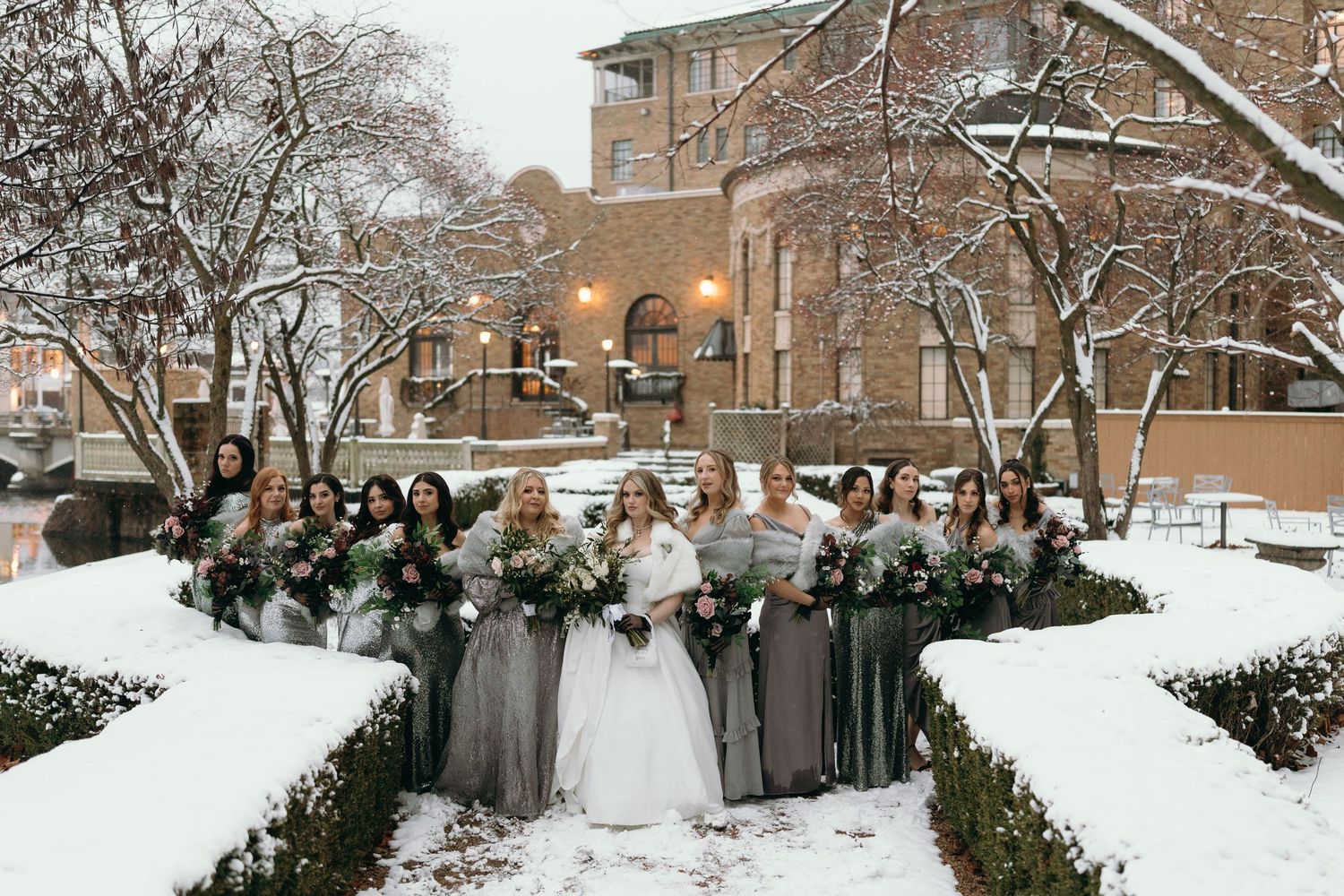 Wedding party in gray dresses and white gown gathered in snow-covered garden.