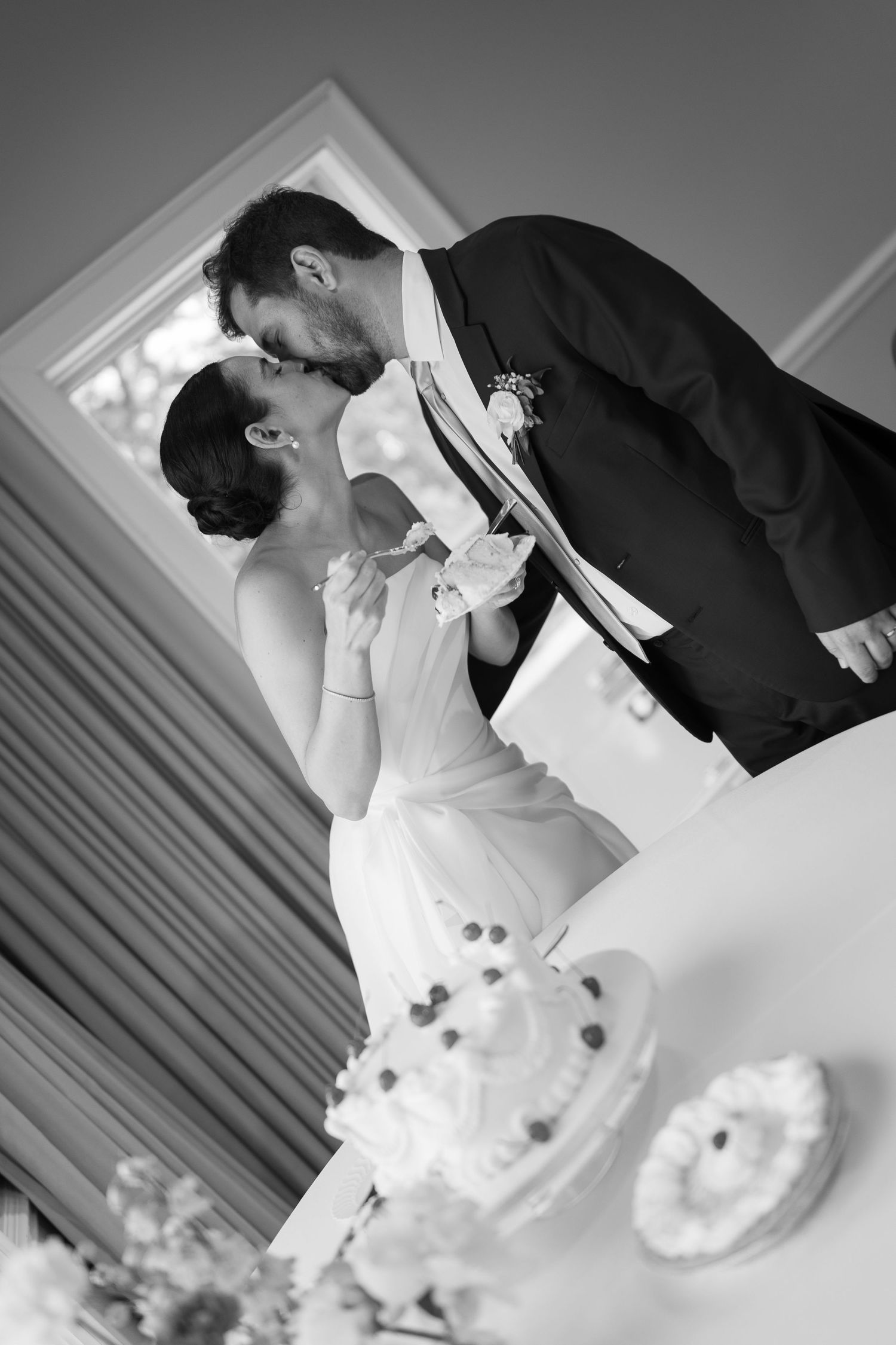 A romantic black and white wedding photo shows a bride and groom sharing a tender kiss behind their wedding cake.