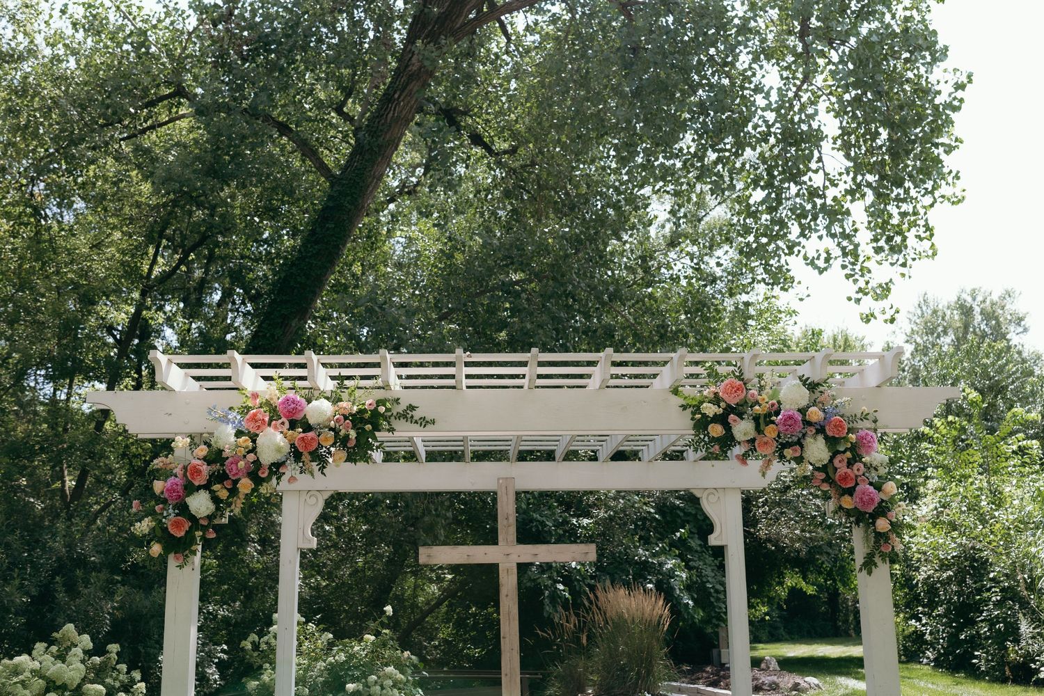 White pergola adorned with pink and white floral arrangements in a lush garden wedding ceremony setting.