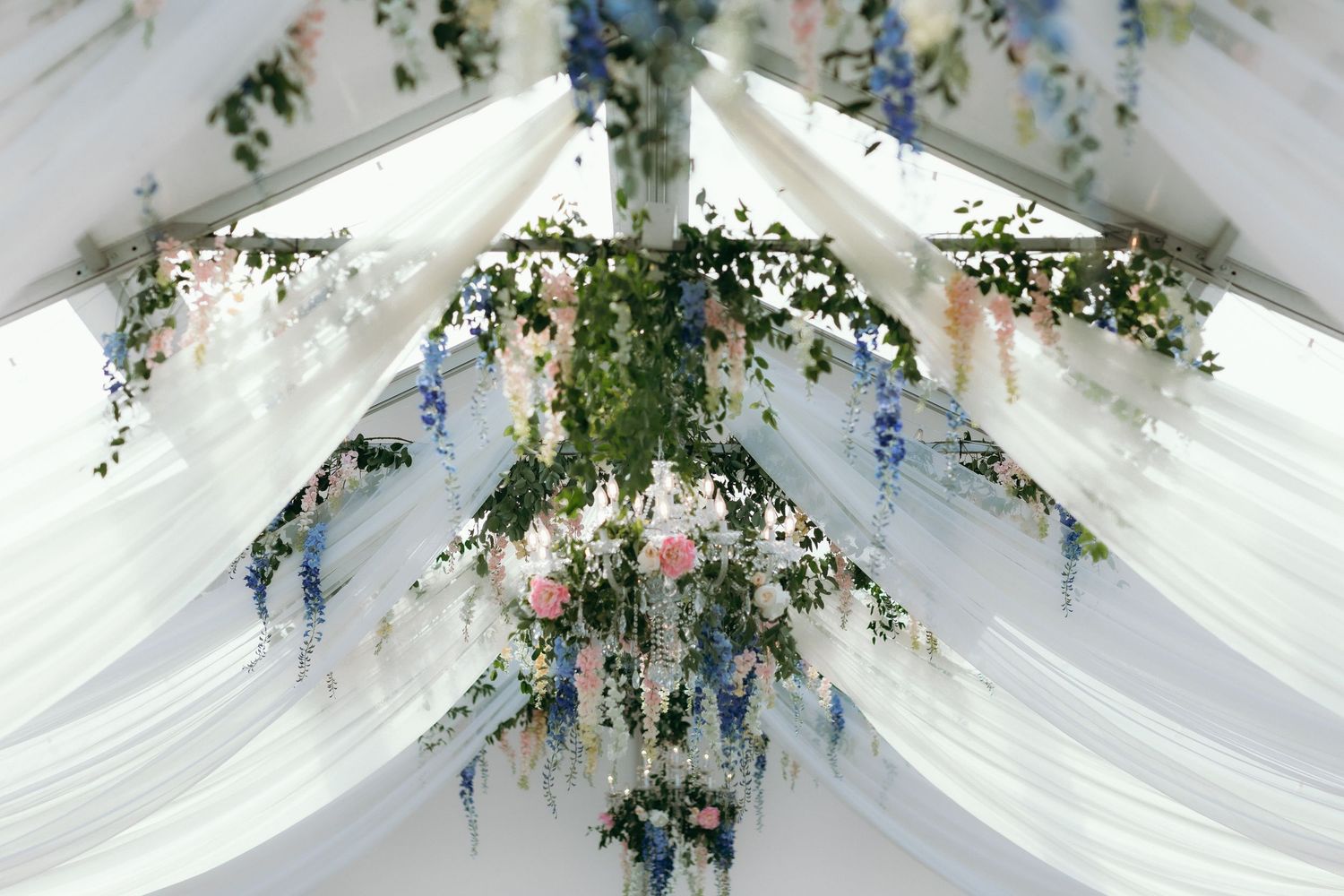 Elegant wedding tent ceiling with white draping, floral garlands of pink roses, blue flowers, and cascading greenery.