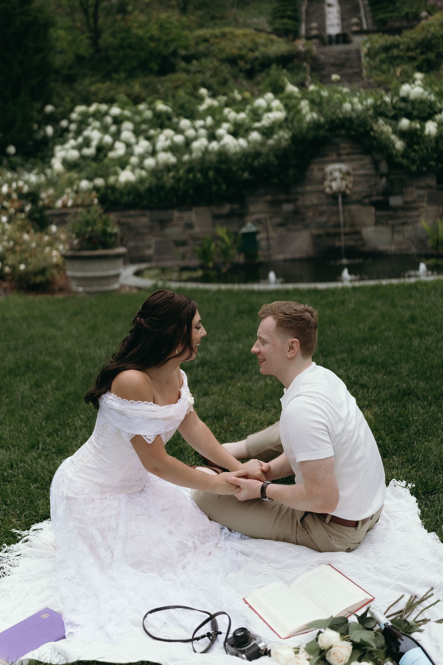 A couple in white outfits shares a tender moment while sitting on a blanket at a garden picnic.