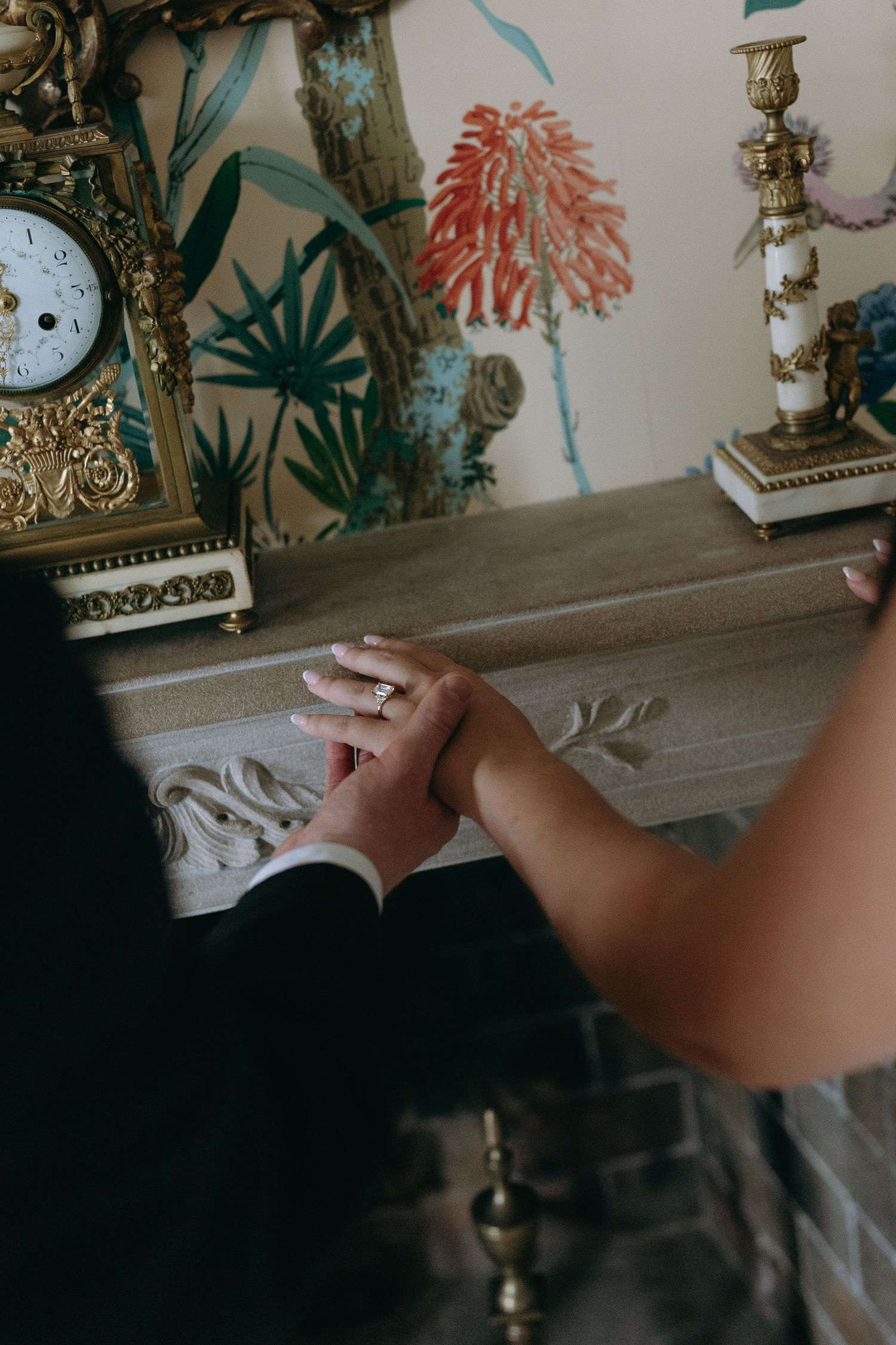 Hand reaching towards a decorative brass clock on a mantle with floral wall art in teal and coral colors in background.
