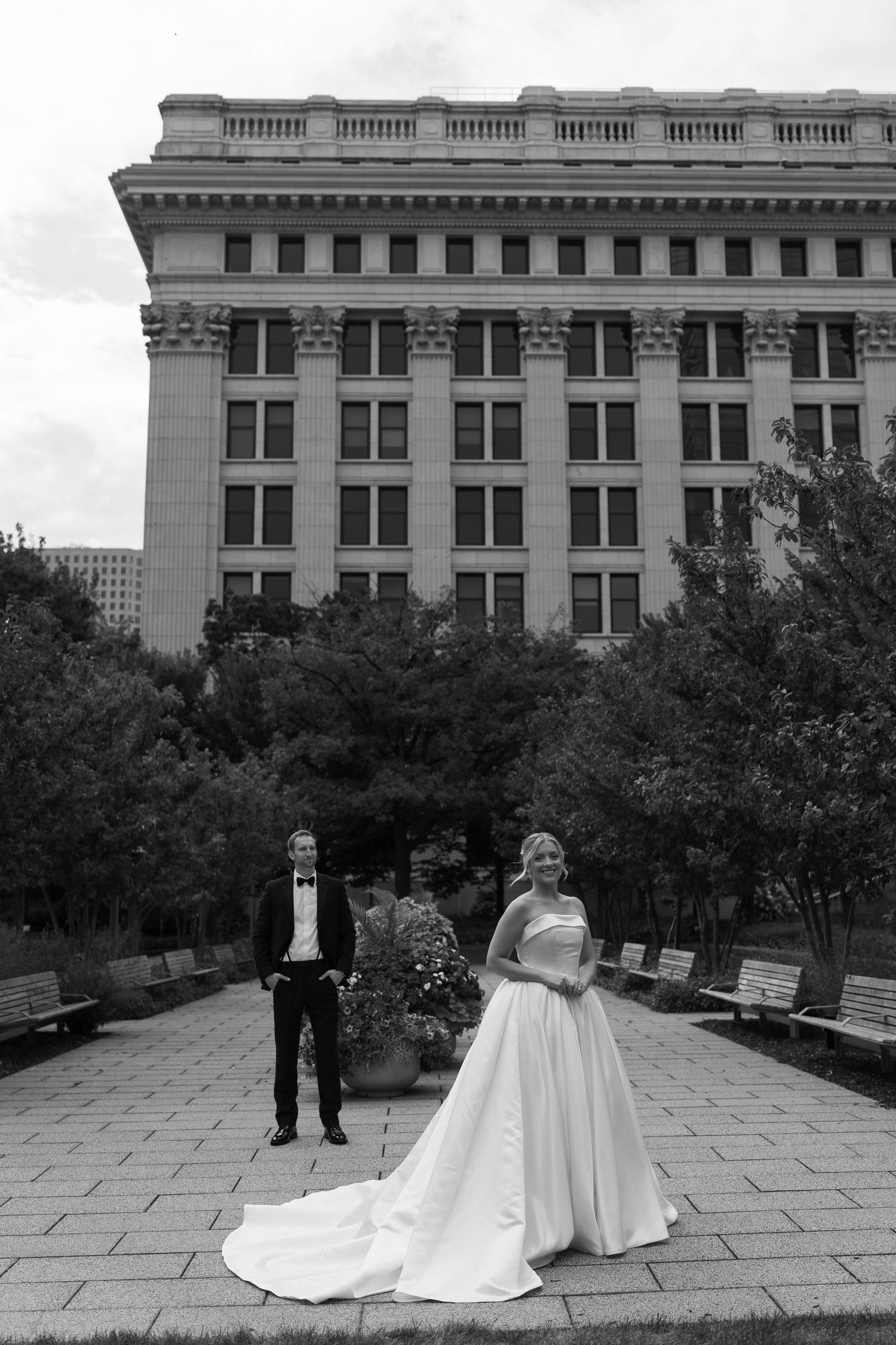 A couple in formal wedding attire poses in front of a historic building with benches and trees in the foreground.