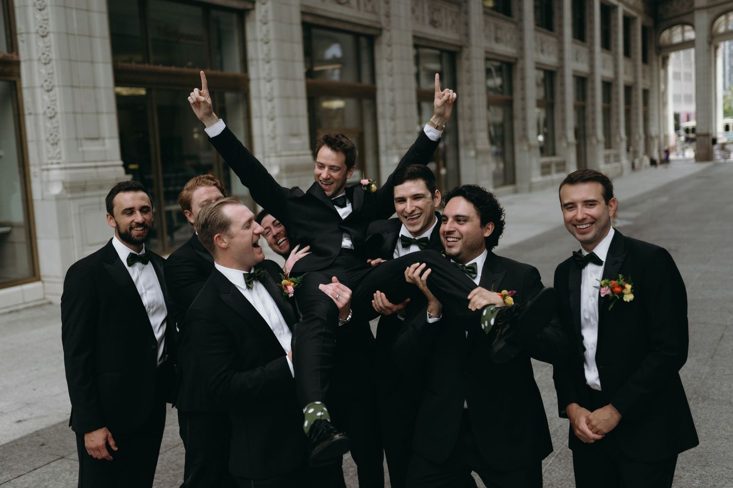 A group of groomsmen in black tuxedos laugh and smile together during a formal wedding event.