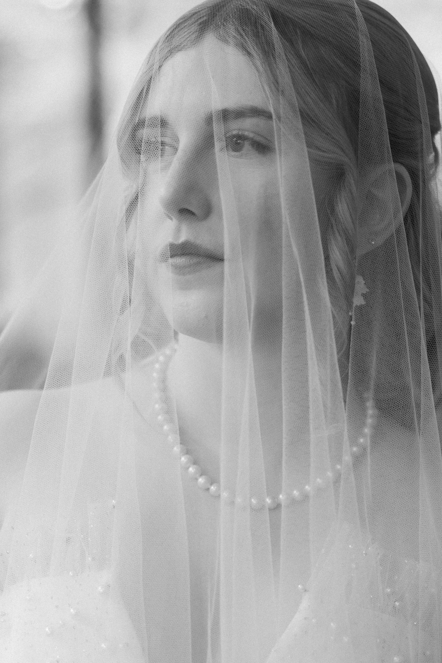Black and white bridal portrait of a young woman wearing a pearl necklace and wedding veil draped over her face.