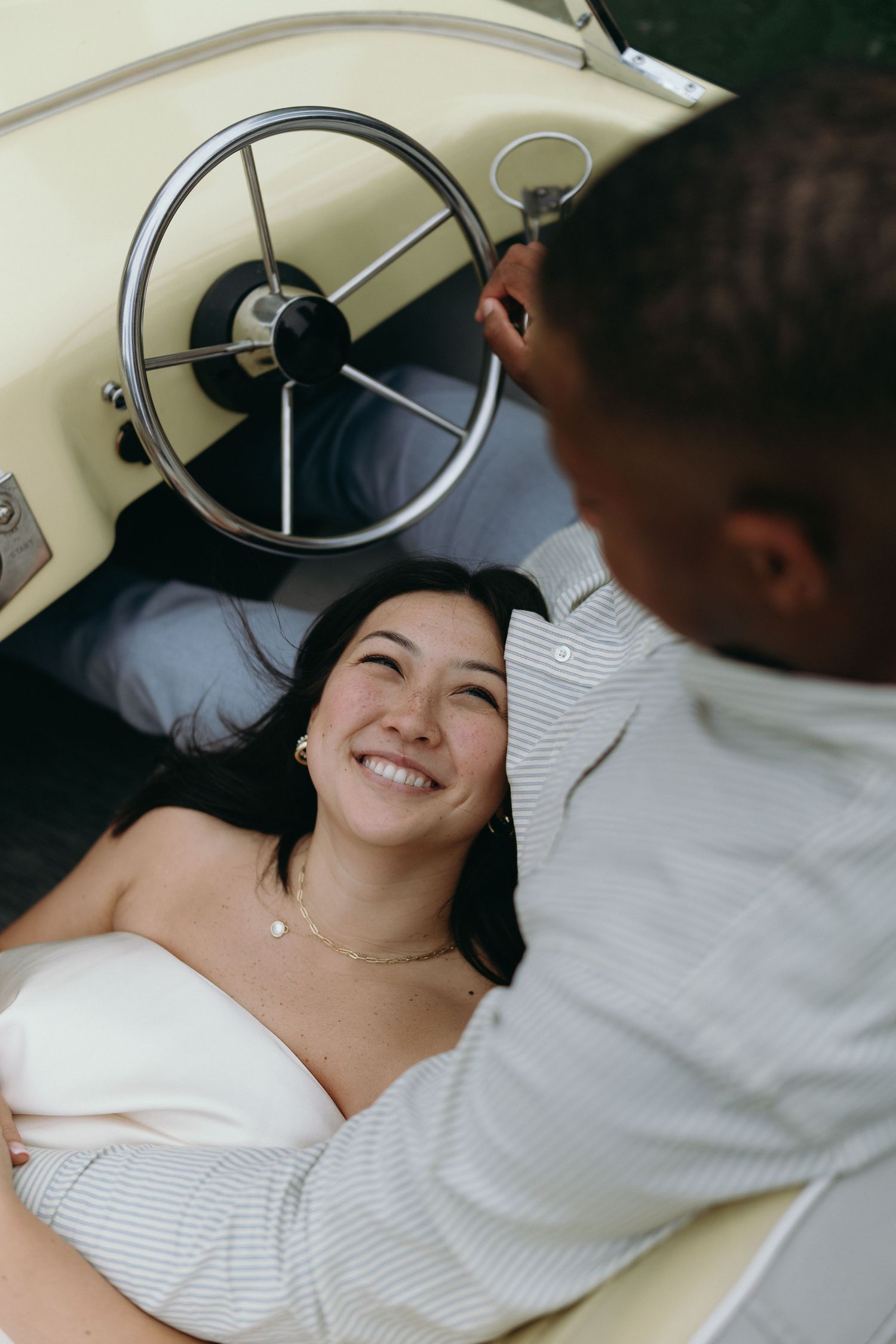 A smiling woman reclines in a vintage cream car while a man in a striped shirt looks down at her tenderly.