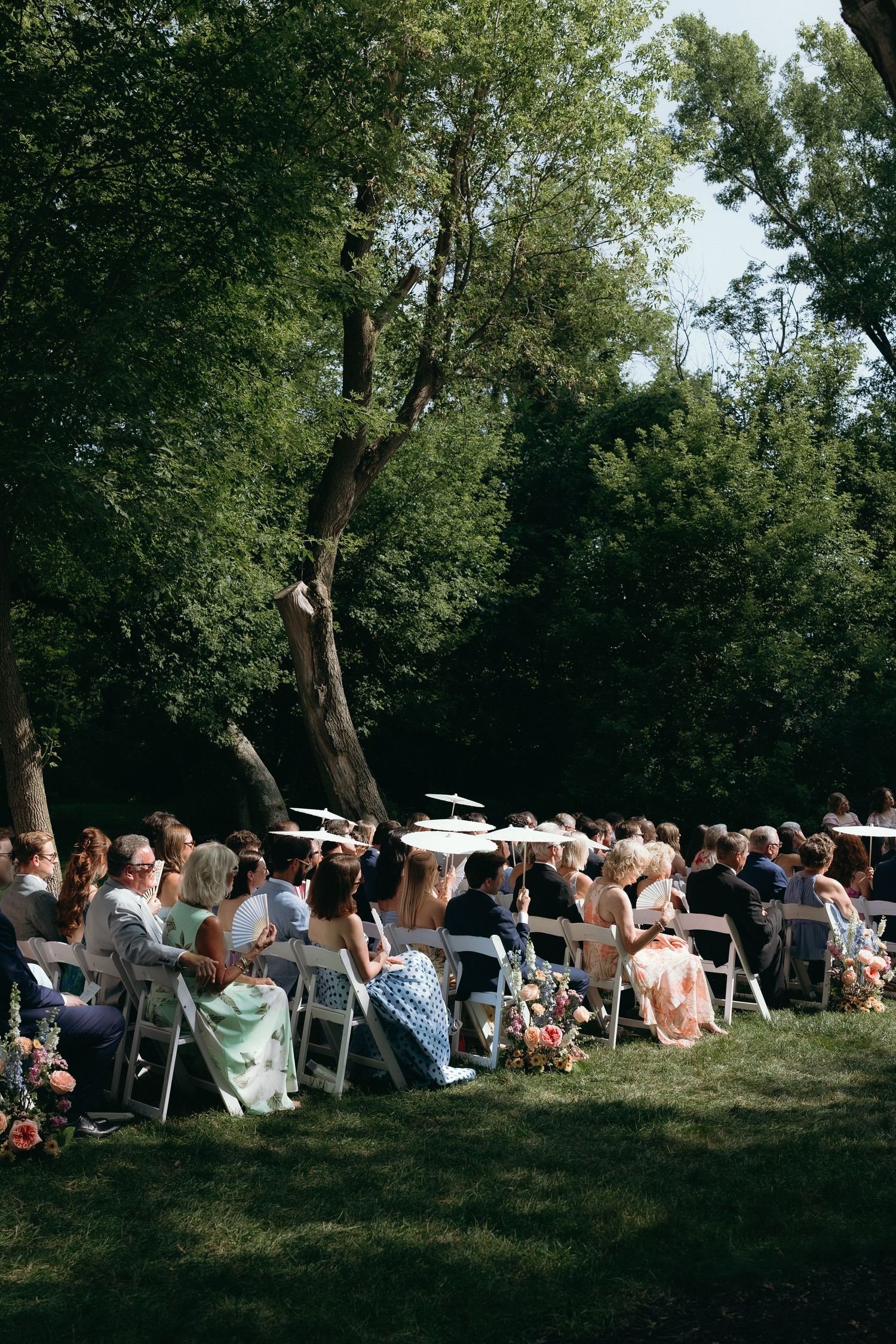 Guests seated in white chairs at an elegant outdoor wedding ceremony surrounded by lush green trees.