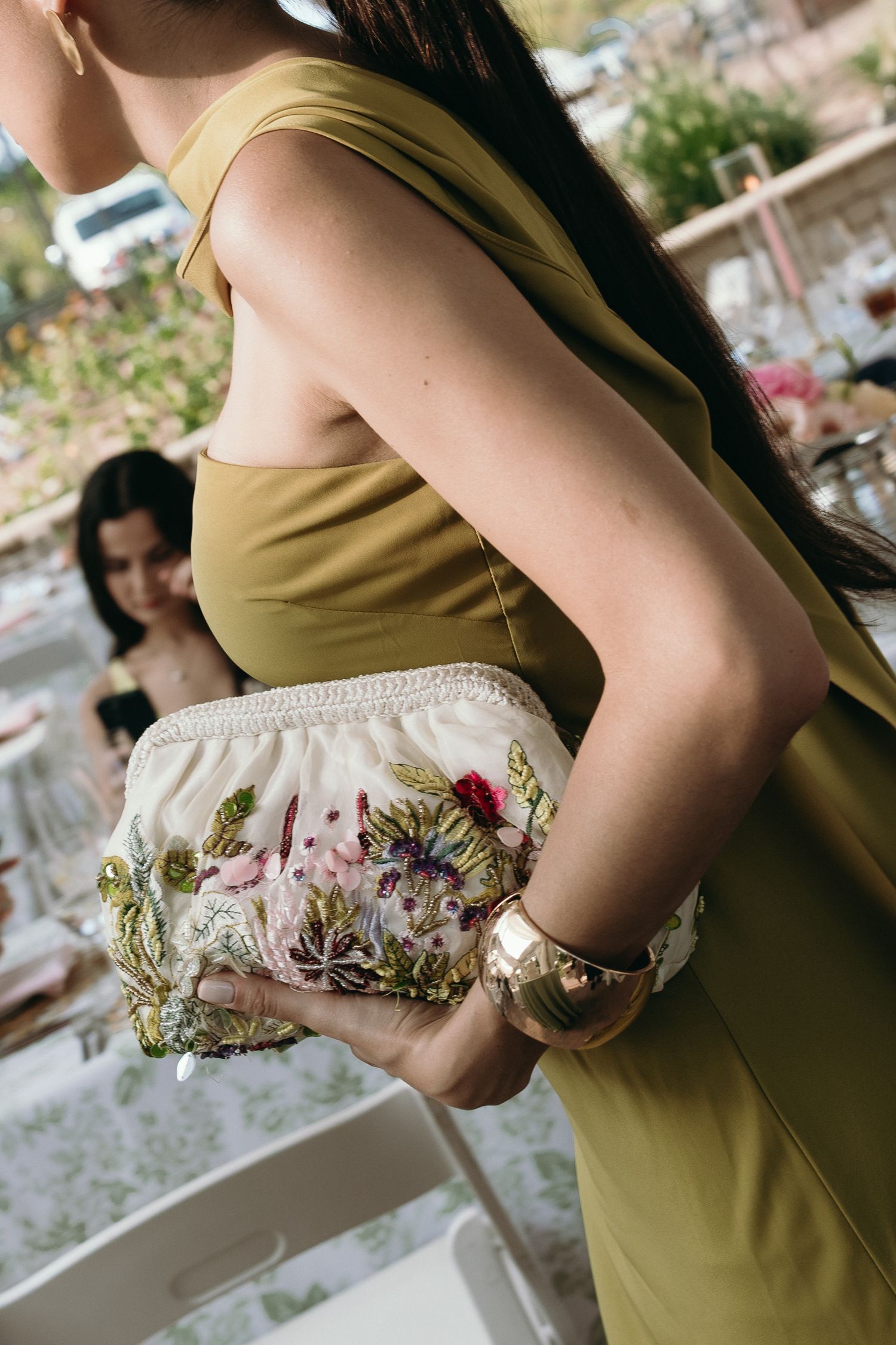 Woman in olive dress holding floral embroidered clutch purse with gold watch at outdoor event.