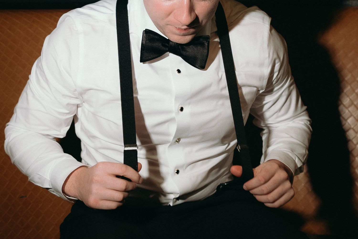 Young groom in white dress shirt, black bow tie, and suspenders getting dressed for a formal wedding ceremony.