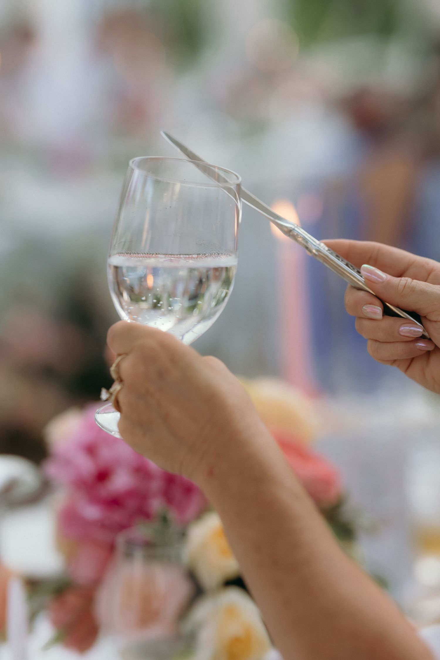 Close-up of hands clinking wine glasses at a wedding reception with colorful floral centerpieces in background.