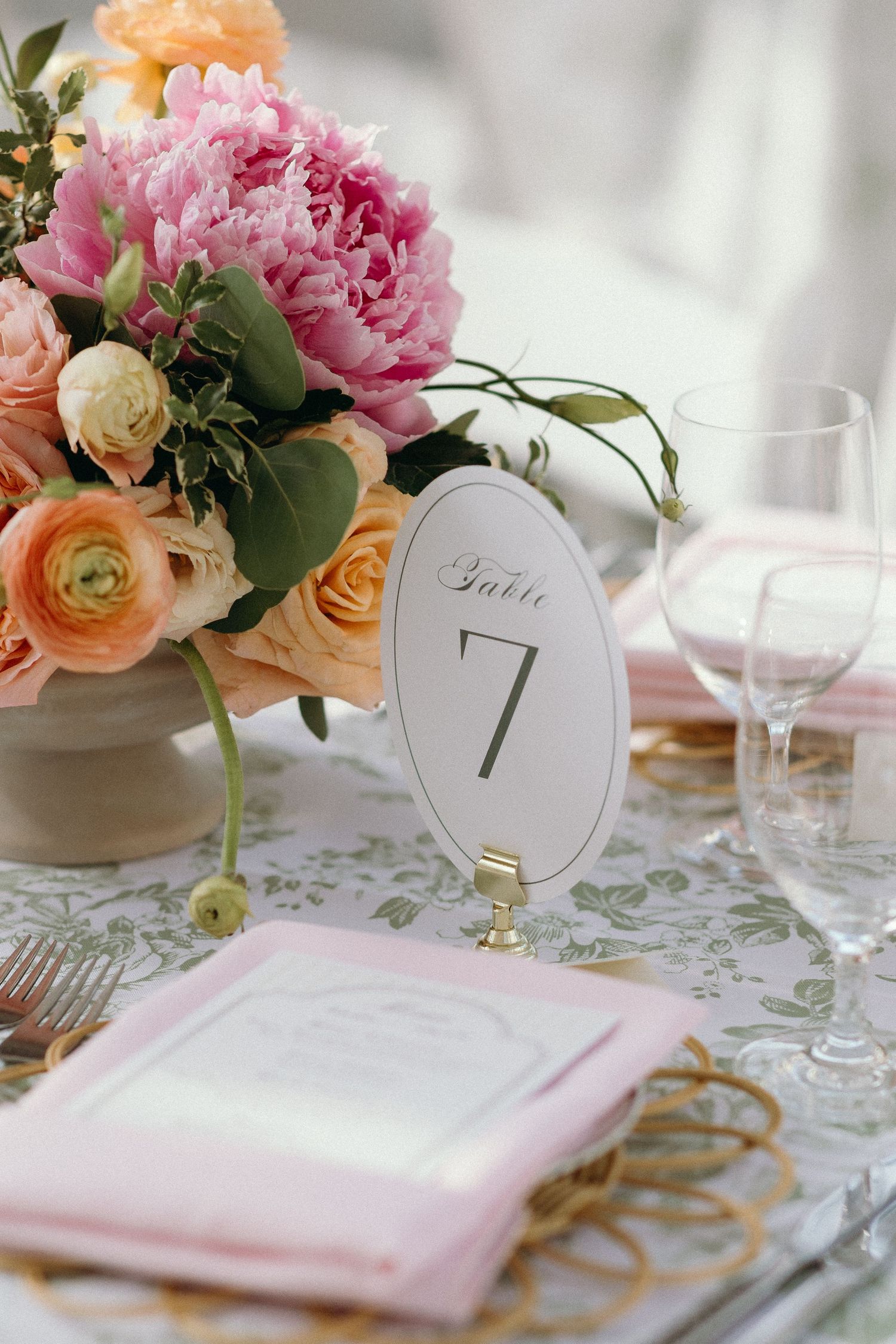 Elegant wedding table setting with pink peonies, peach roses, pink napkins, and a gold table number 7 sign.