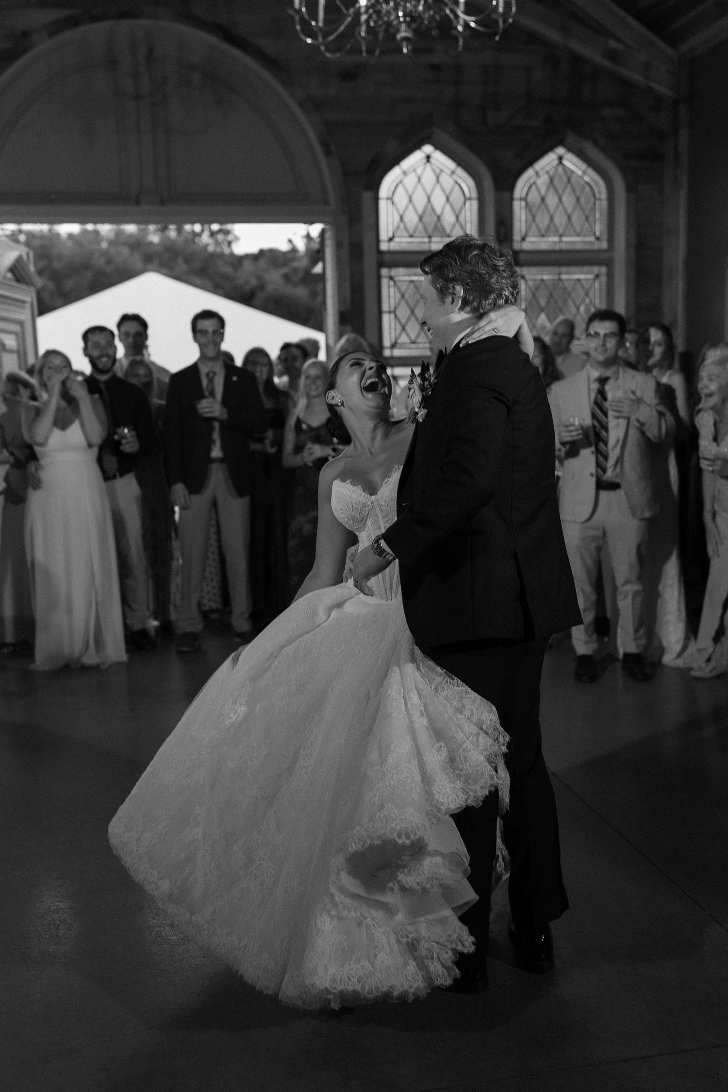 Black and white photo of bride and groom sharing romantic first dance surrounded by wedding guests.