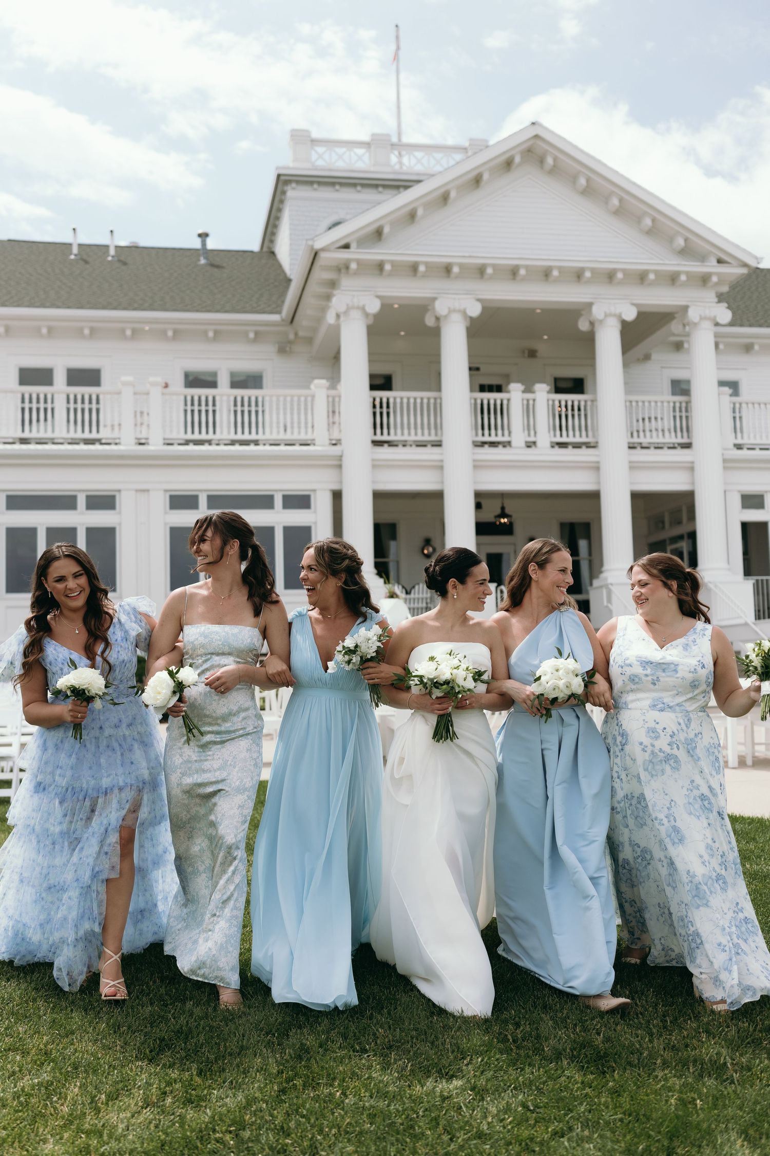 Bridal party in light blue and floral dresses poses together on lawn in front of elegant white colonial mansion.