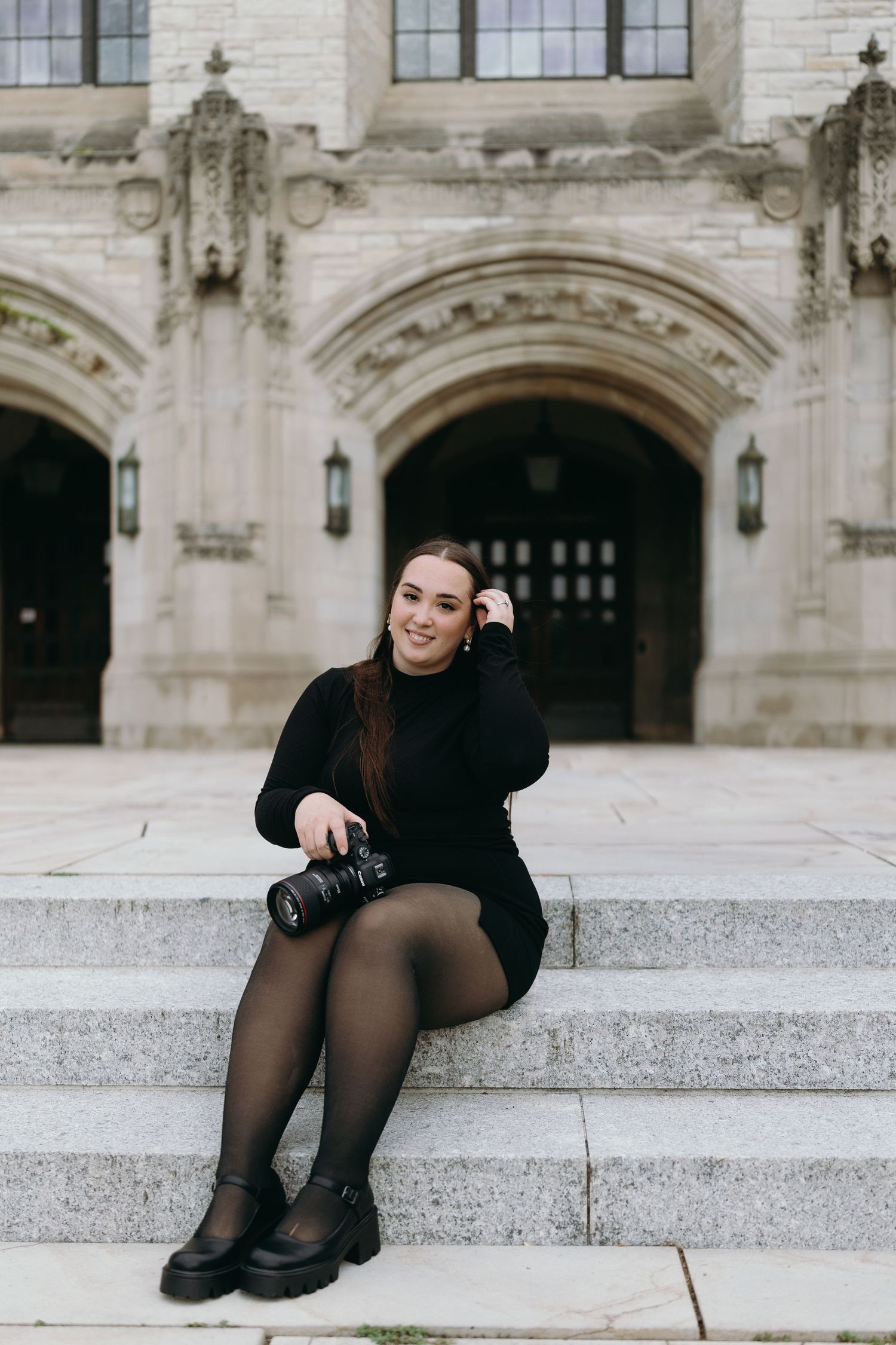 Photographer in black dress sits on stone steps of historic building with ornate architecture.