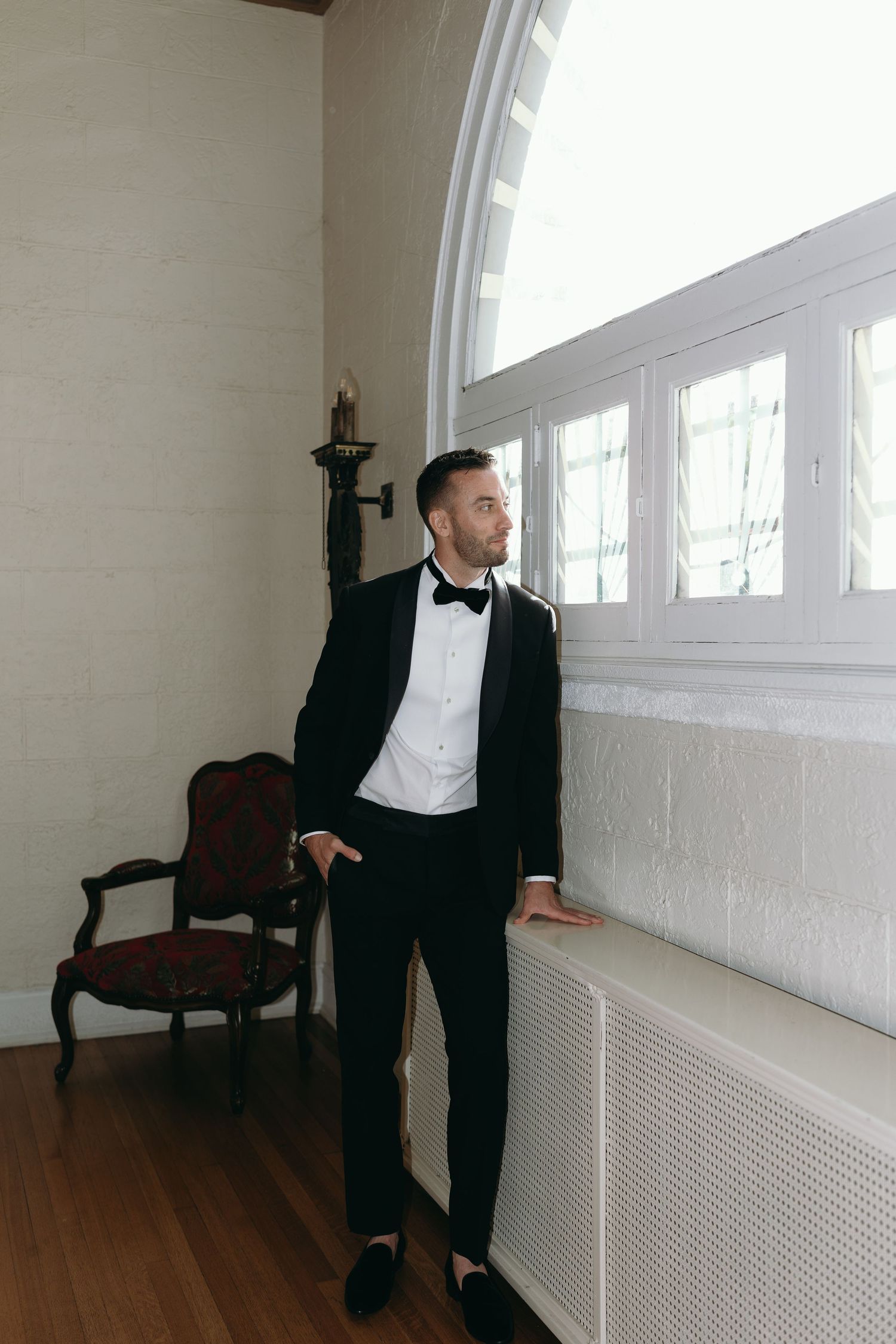 A classic tuxedo-clad figure stands beside a window in an elegant room with hardwood floors and vintage furniture.