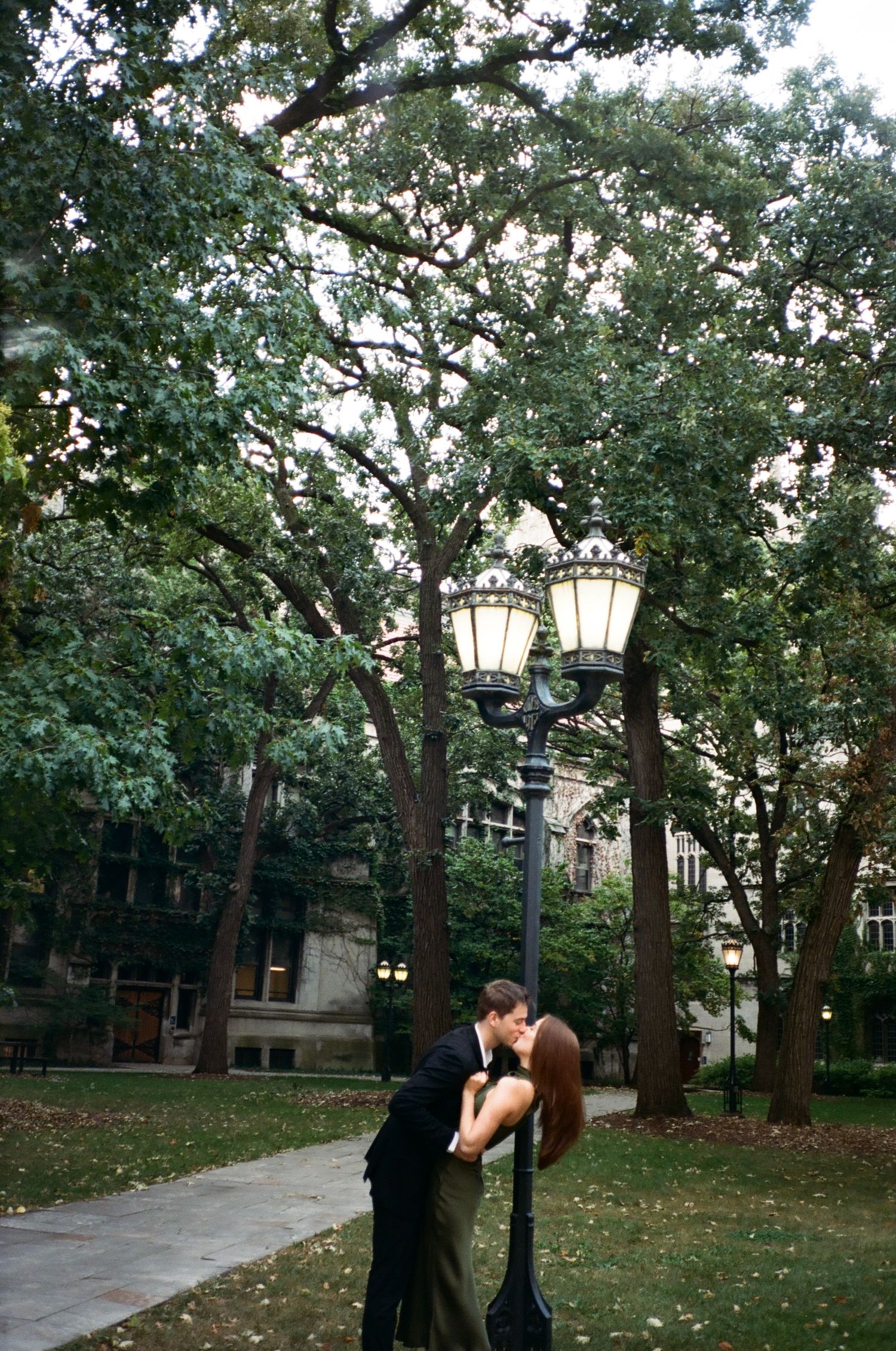 Couple kissing under a vintage street lamp in a lush green park with tall trees and historic building.