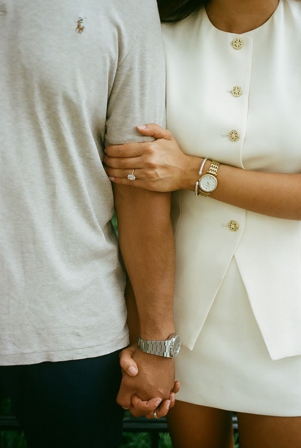 Couple holding hands, woman wearing diamond ring, gold watch, bracelet, white blazer dress with gold buttons.