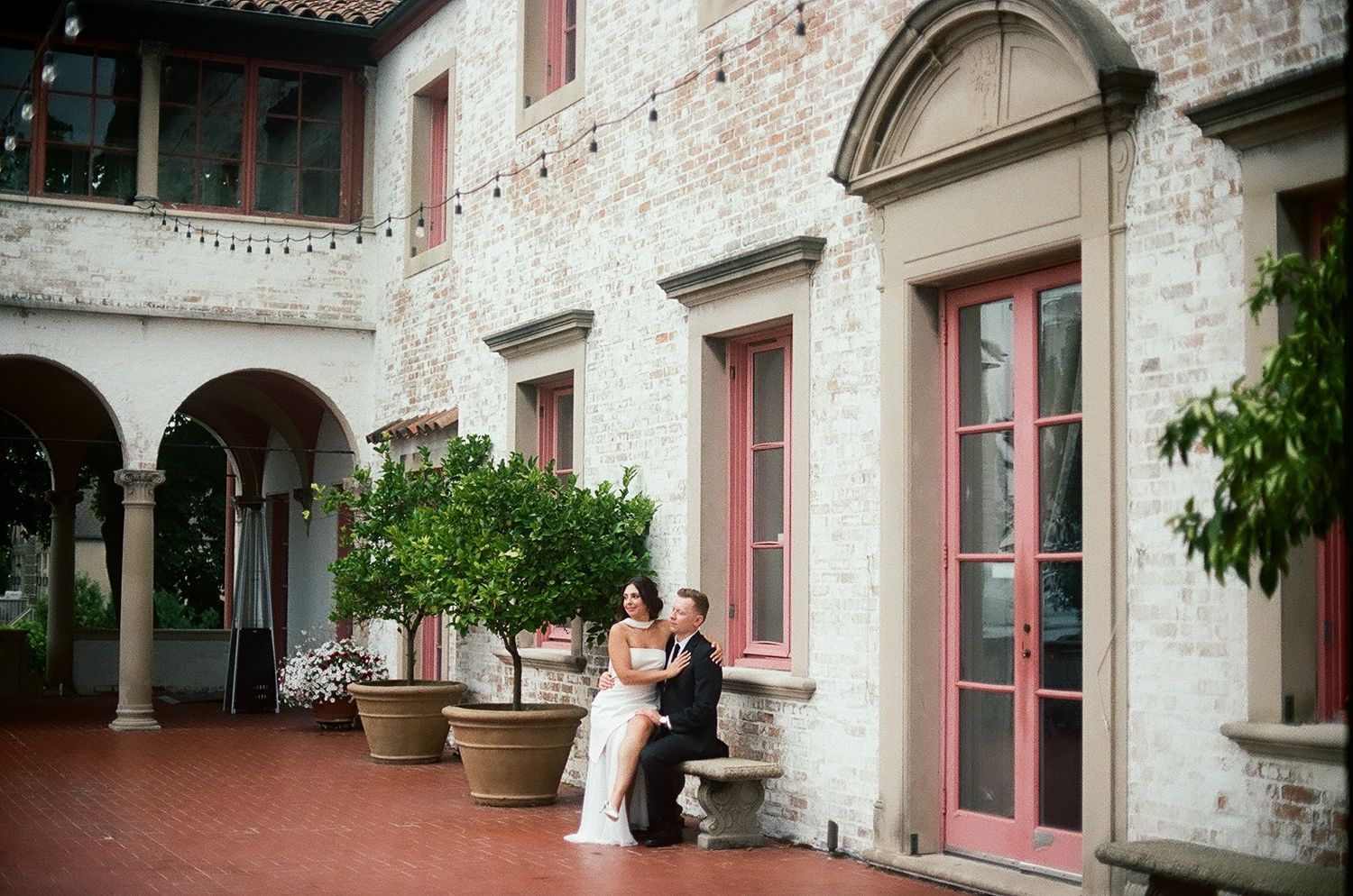 Bride and groom share a romantic moment in an elegant courtyard with arched walkways and potted trees.