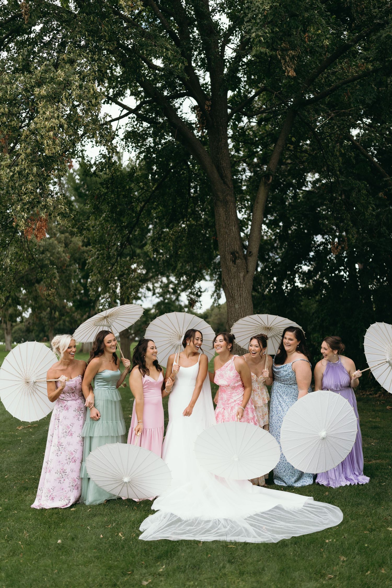 Wedding party lounging on grass with white parasols in a casual outdoor photo session.