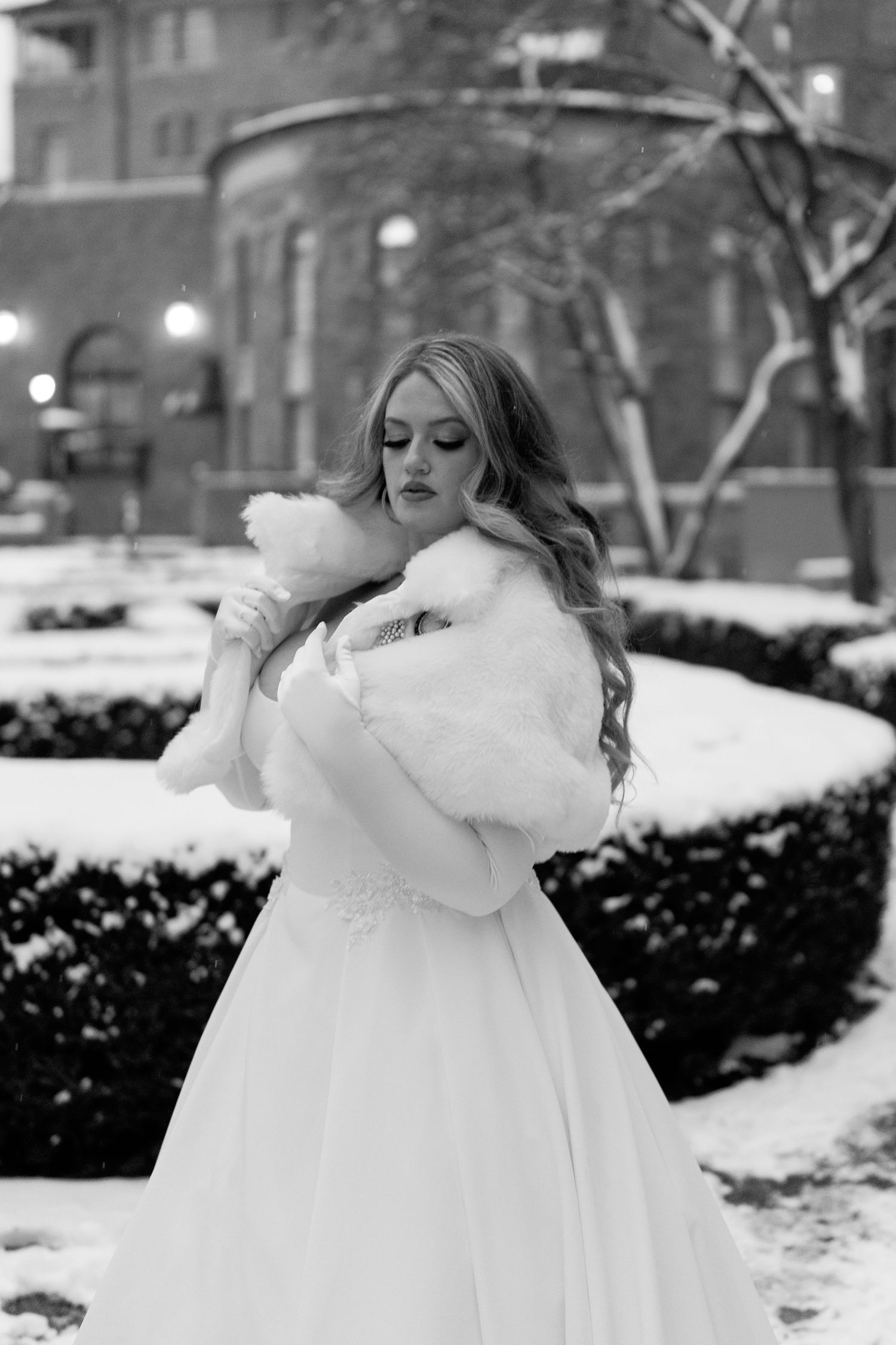 Winter bride in white gown and fur wrap poses elegantly in snowy courtyard.