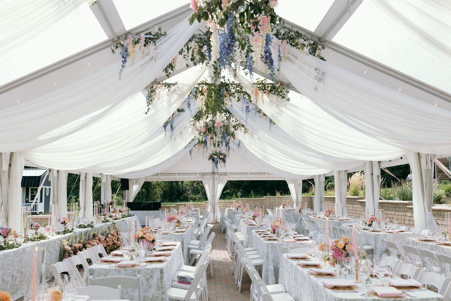 Stunning wedding reception tent with white draping, hanging floral installations, and elegantly set round tables.