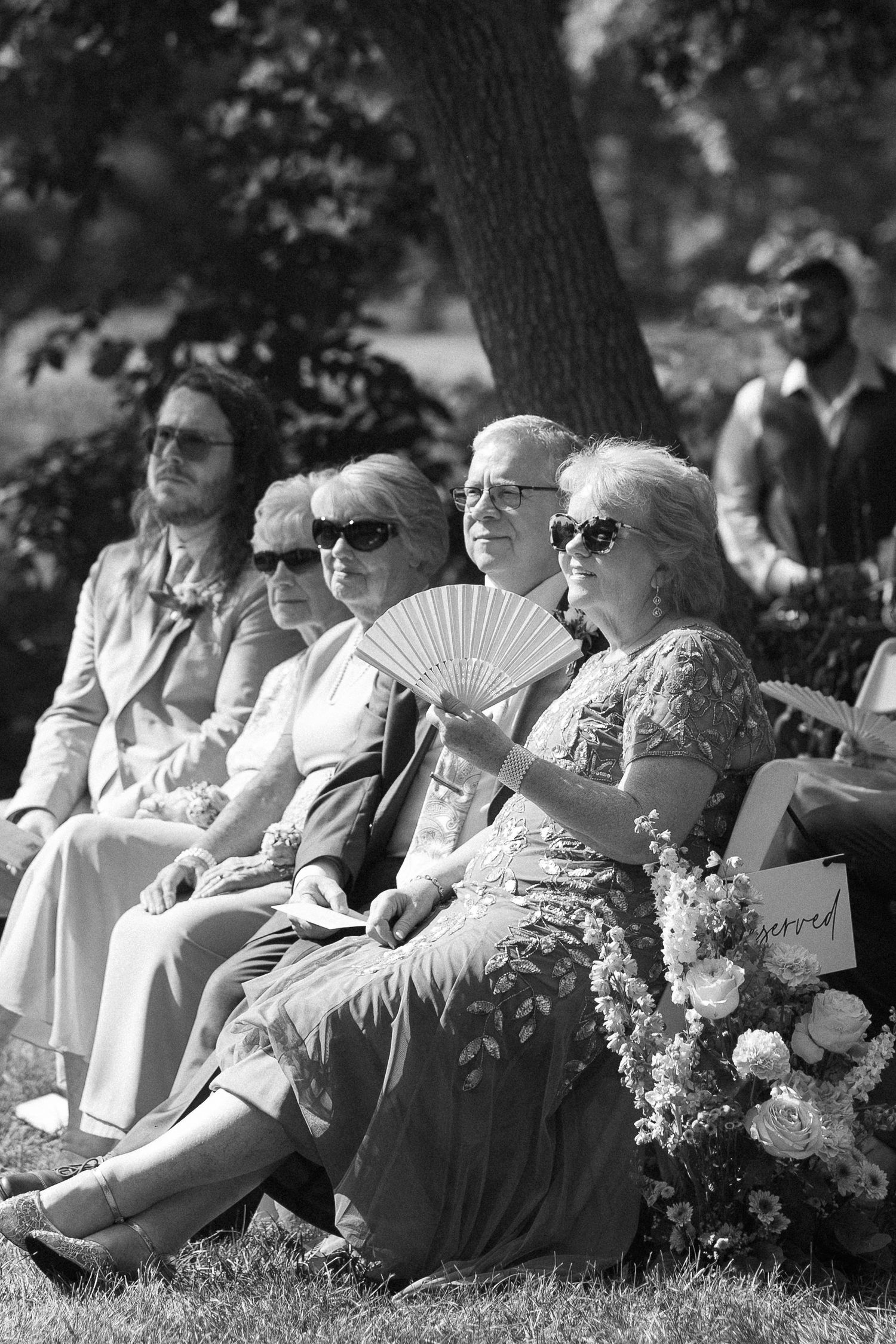 Black and white photo of wedding guests seated outdoors, with a woman using a hand fan to stay cool.