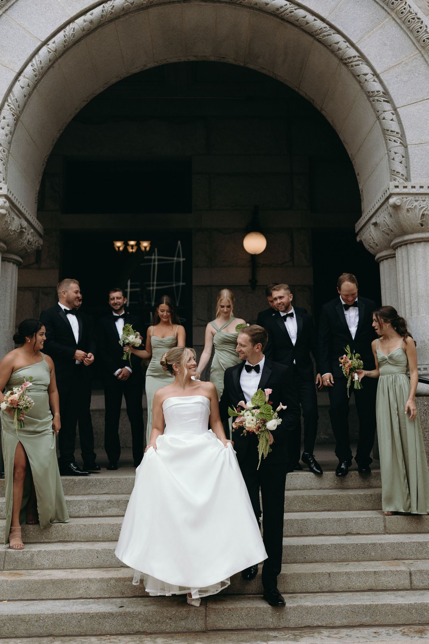 Wedding party in formal attire poses on grand stone steps of an elegant arched building.