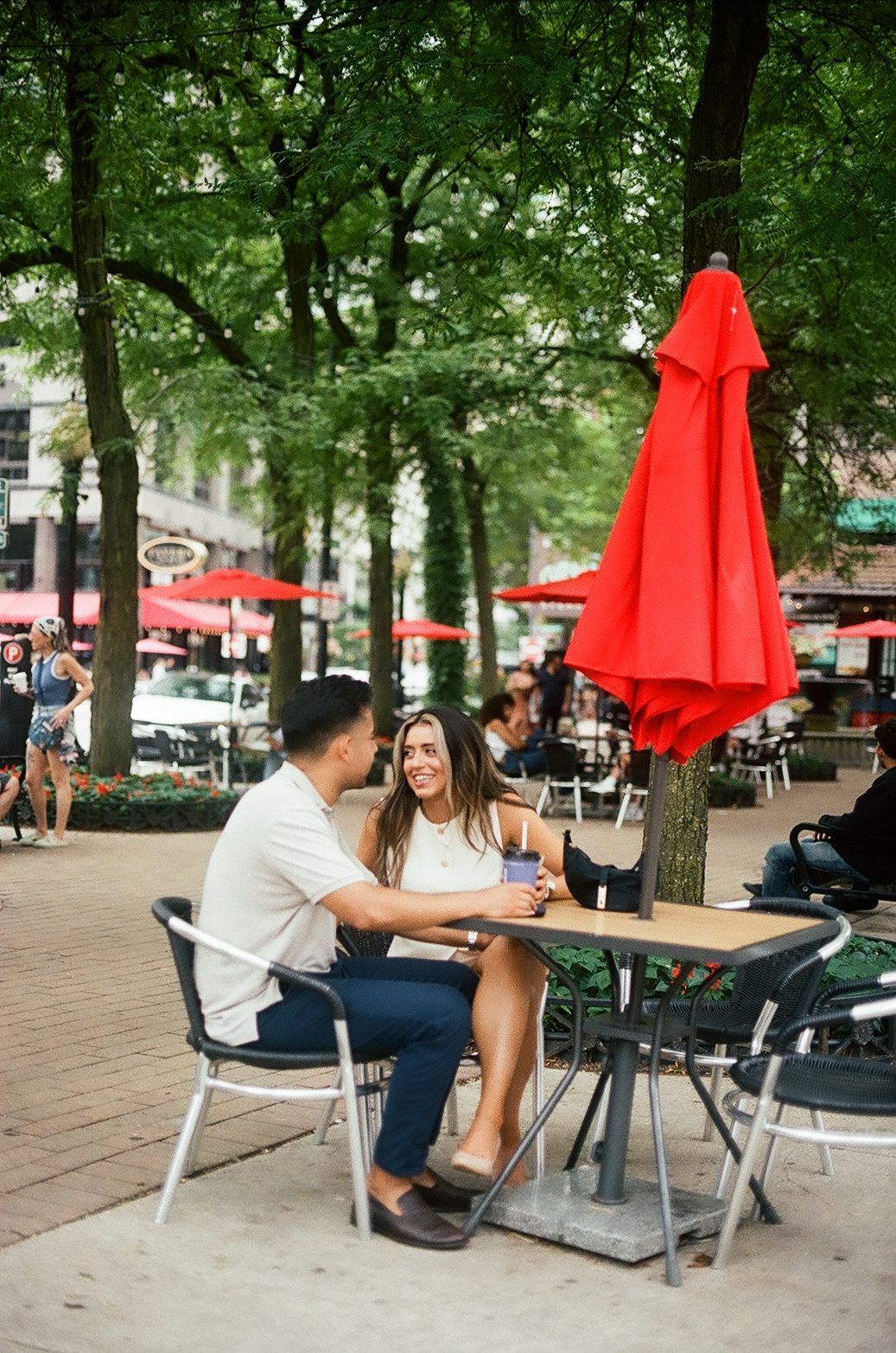 A couple sits at an outdoor cafe table with red umbrellas in a tree-lined urban setting.