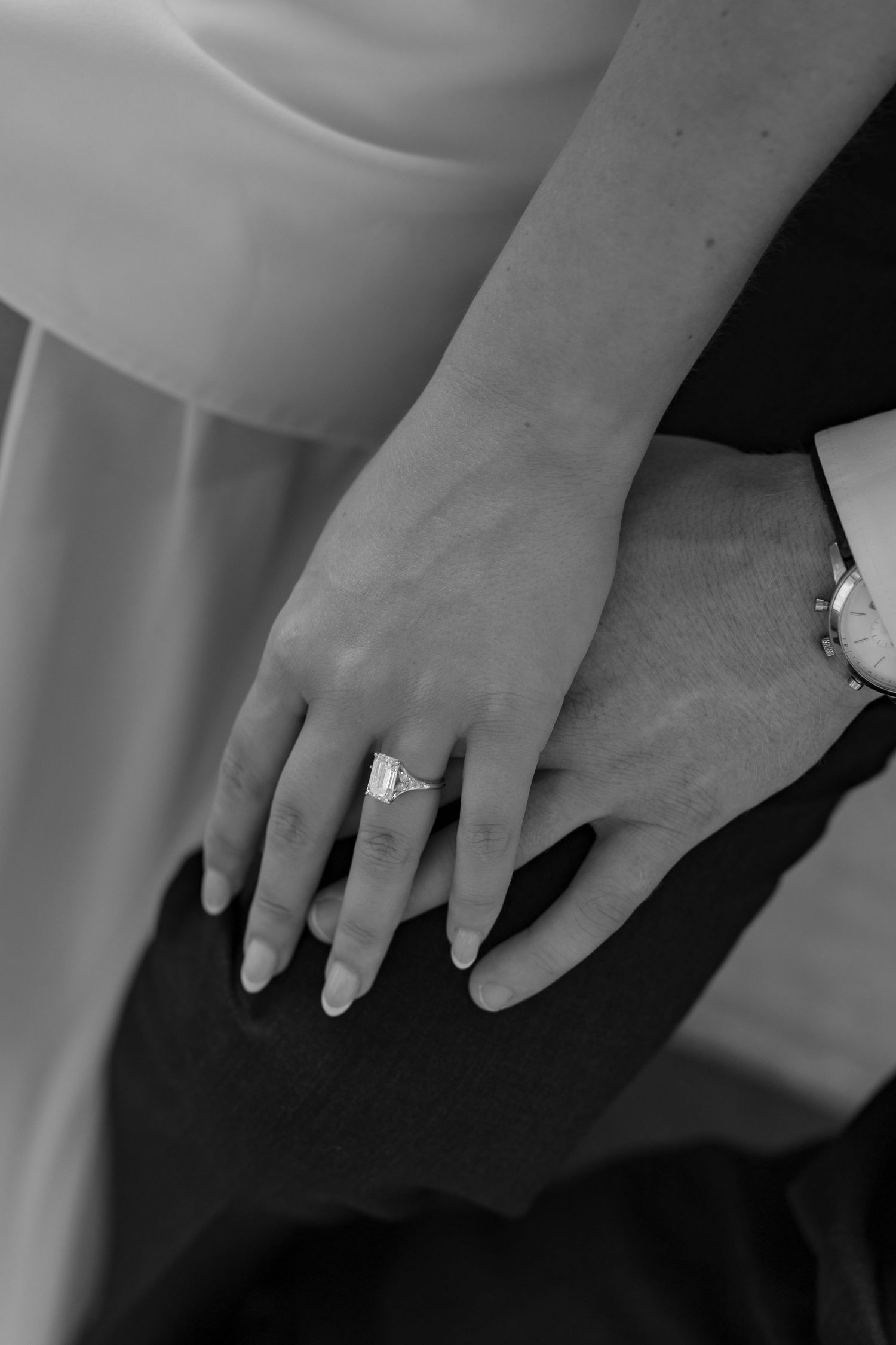A black and white close-up photograph of hands wearing an engagement ring with a white dress in the background.