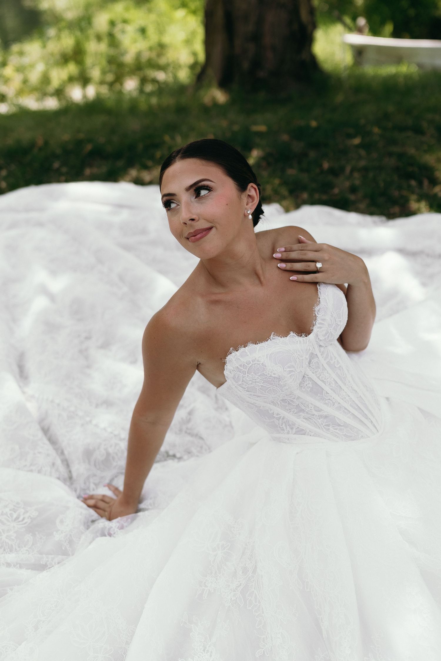 Bride in strapless white lace ball gown sitting outdoors on grass, smiling elegantly with hand on shoulder.