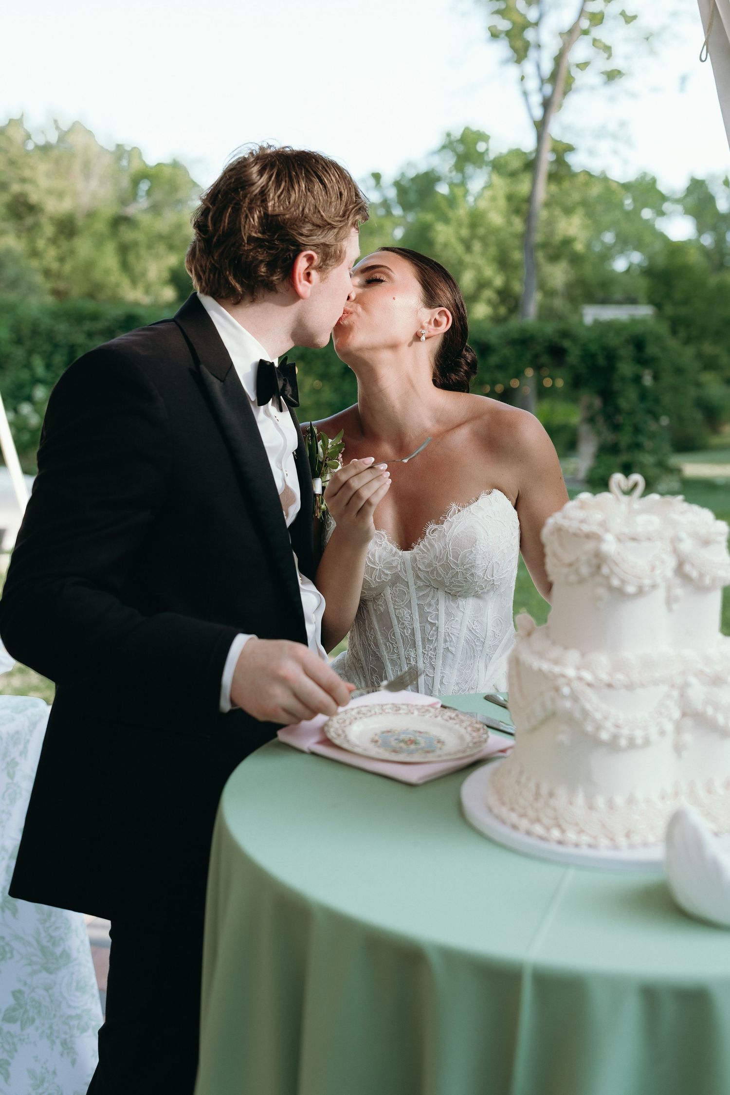 Bride and groom share a kiss while cutting their elegant white tiered wedding cake outdoors.