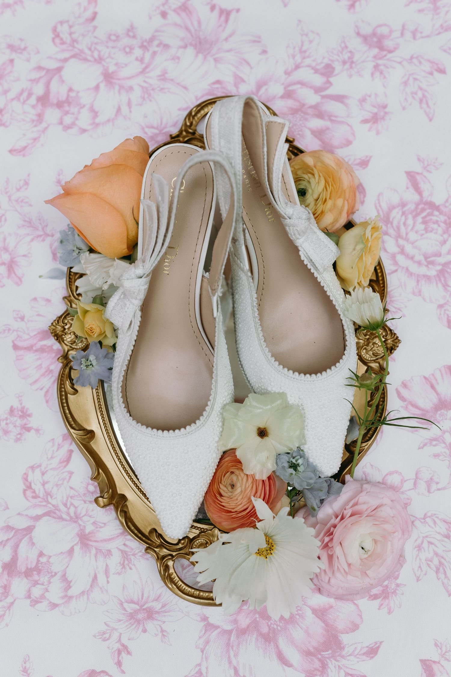 White bridal heels displayed on a gold tray surrounded by peach, white, and blue flowers on a floral pink background.