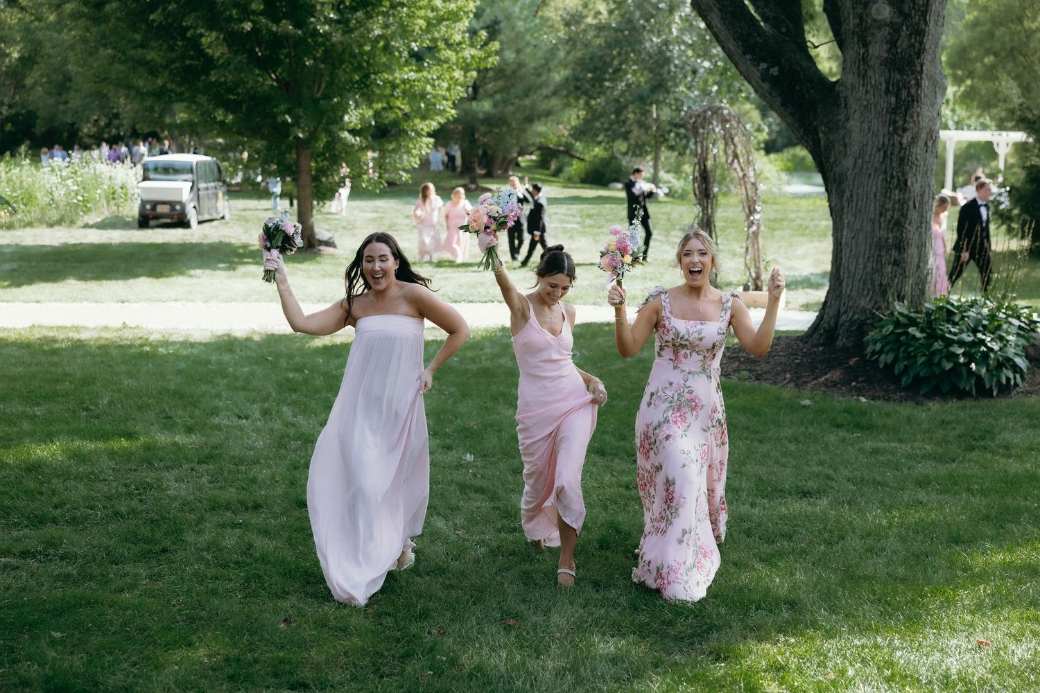 Three bridesmaids in pink dresses walk joyfully across green lawn at outdoor wedding celebration.