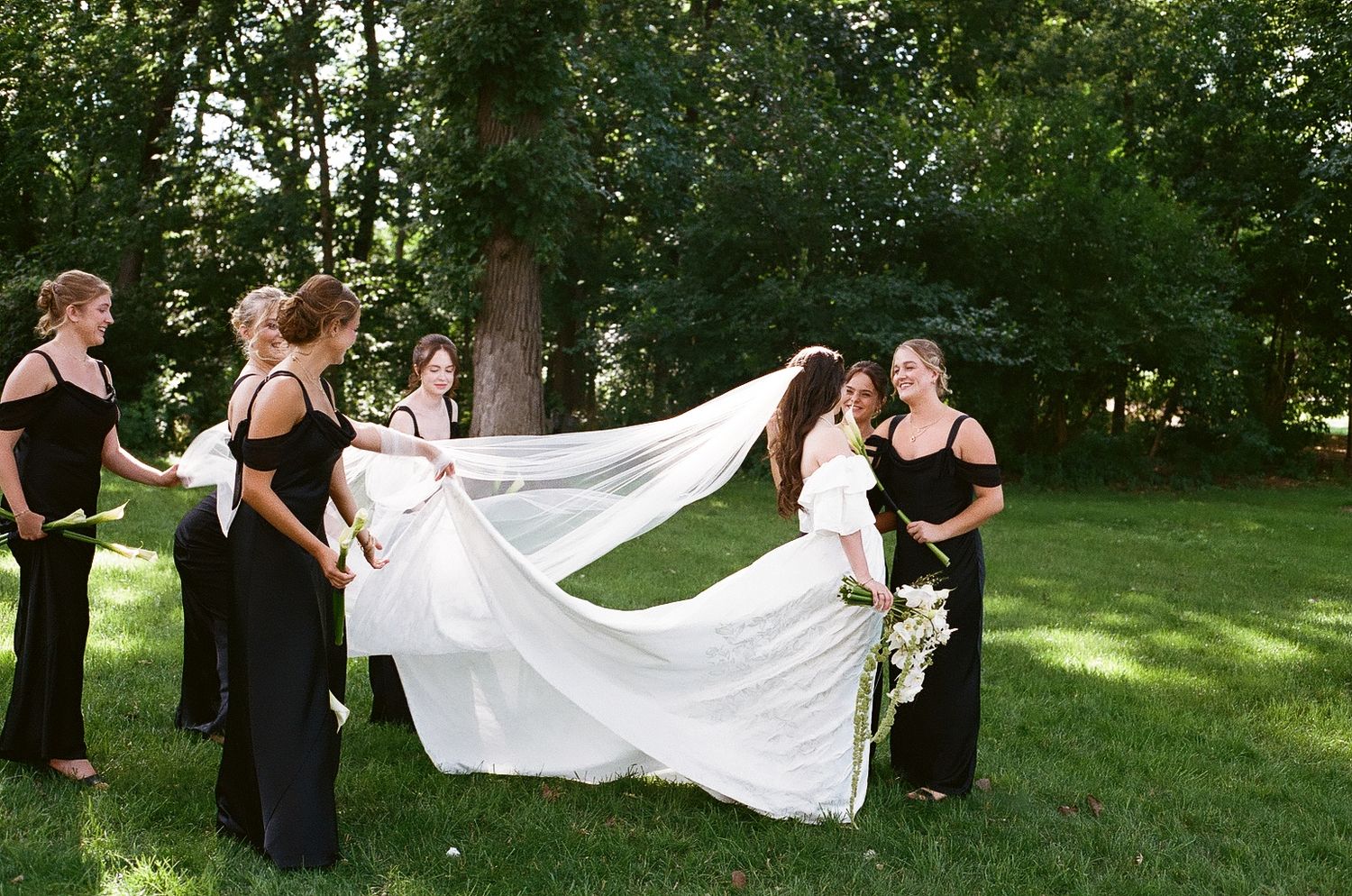 Bridesmaids in black dresses help spread a bride's flowing white wedding gown and veil on a lush green lawn.