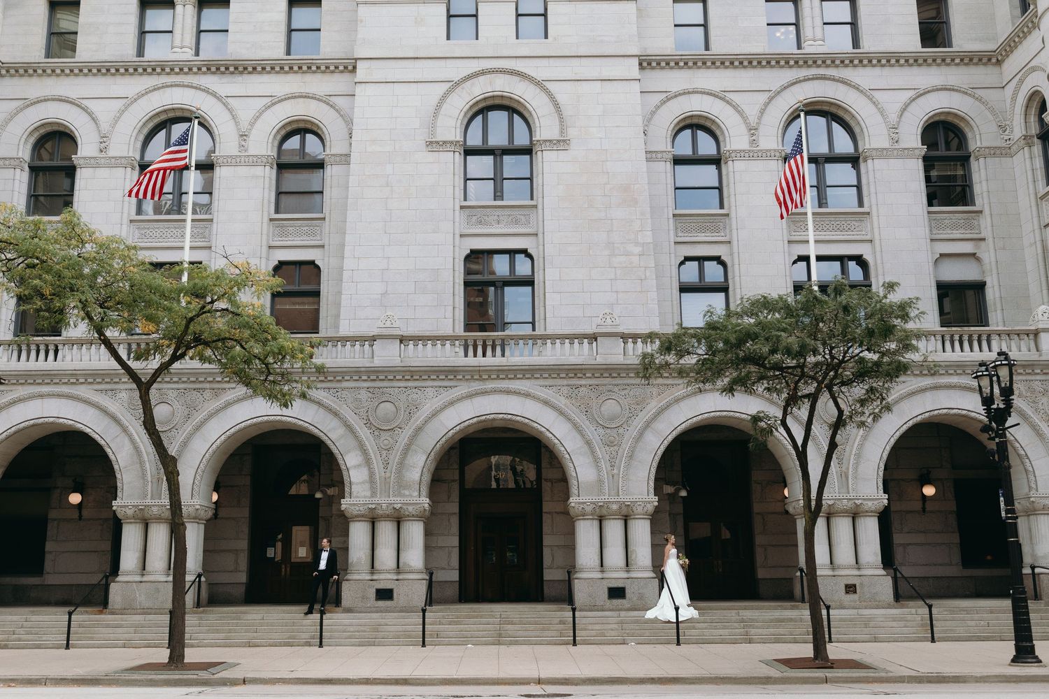 Elegant stone building facade with arched windows, American flags, and trees along a city sidewalk.