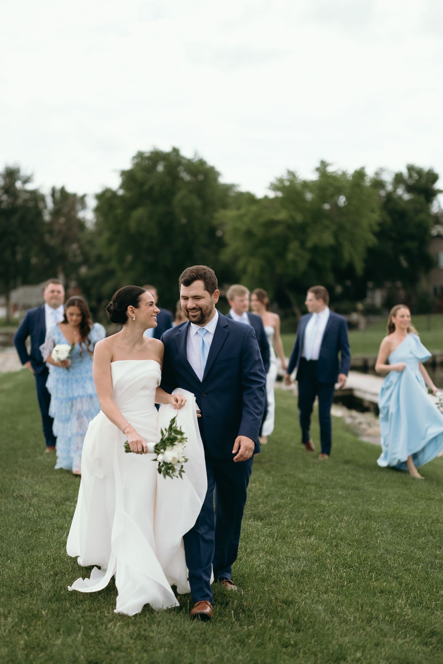 Wedding party walks across a green lawn with the bride in white and bridesmaids in light blue dresses following behind.