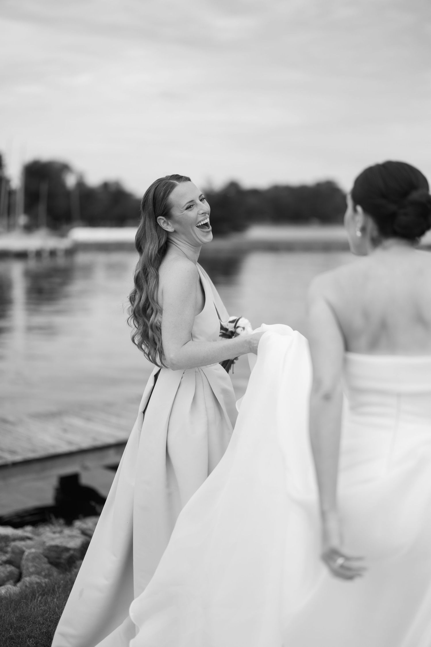 A joyful bride in a white wedding dress laughs candidly while standing on an outdoor deck near the water.