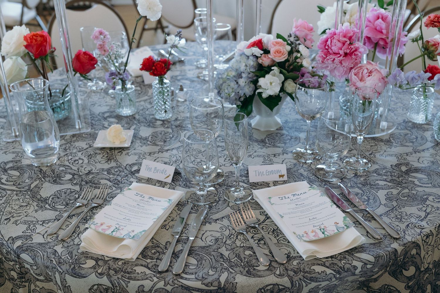 Elegant place settings at a wedding reception table with pink and red roses and crystal stemware reflect in a silver charger plate.