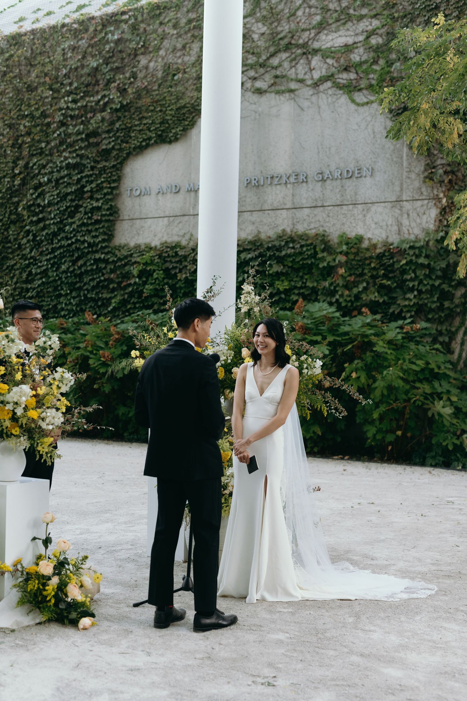 A wedding ceremony takes place against an ivy-covered wall with yellow floral arrangements.